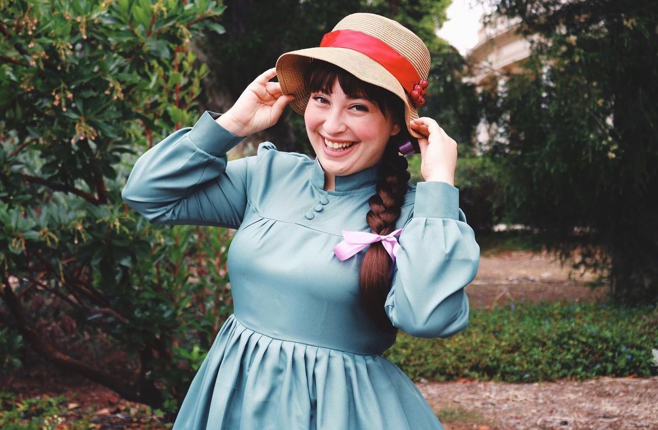 A cosplayer in vintage blue dress and red-banded straw hat poses playfully in a garden.