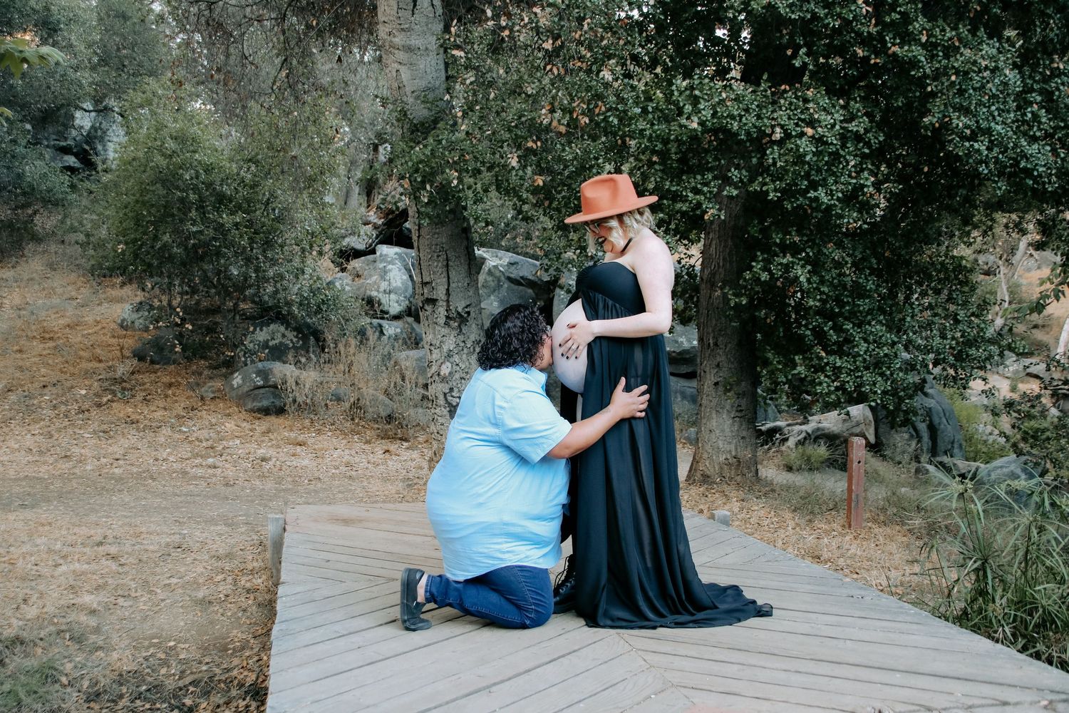 Tender moment captured during pregnancy photoshoot on wooden platform surrounded by trees.