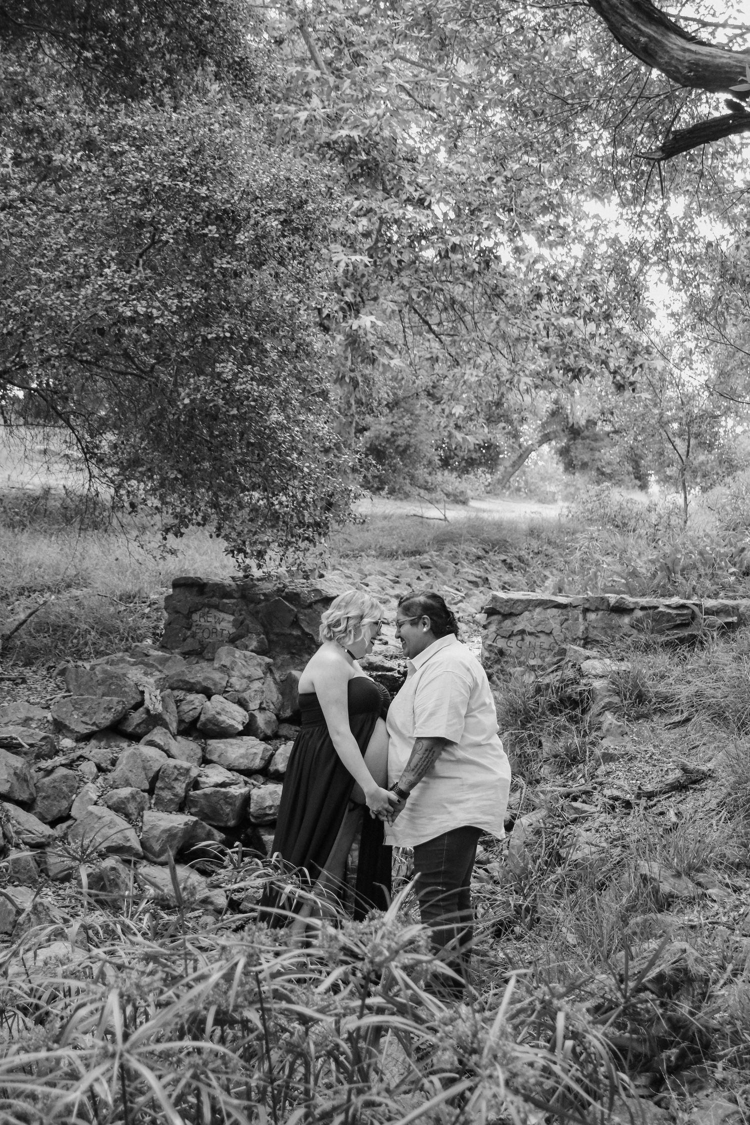 Two people sit together on rocky ground in a wooded outdoor setting, surrounded by trees and vegetation.