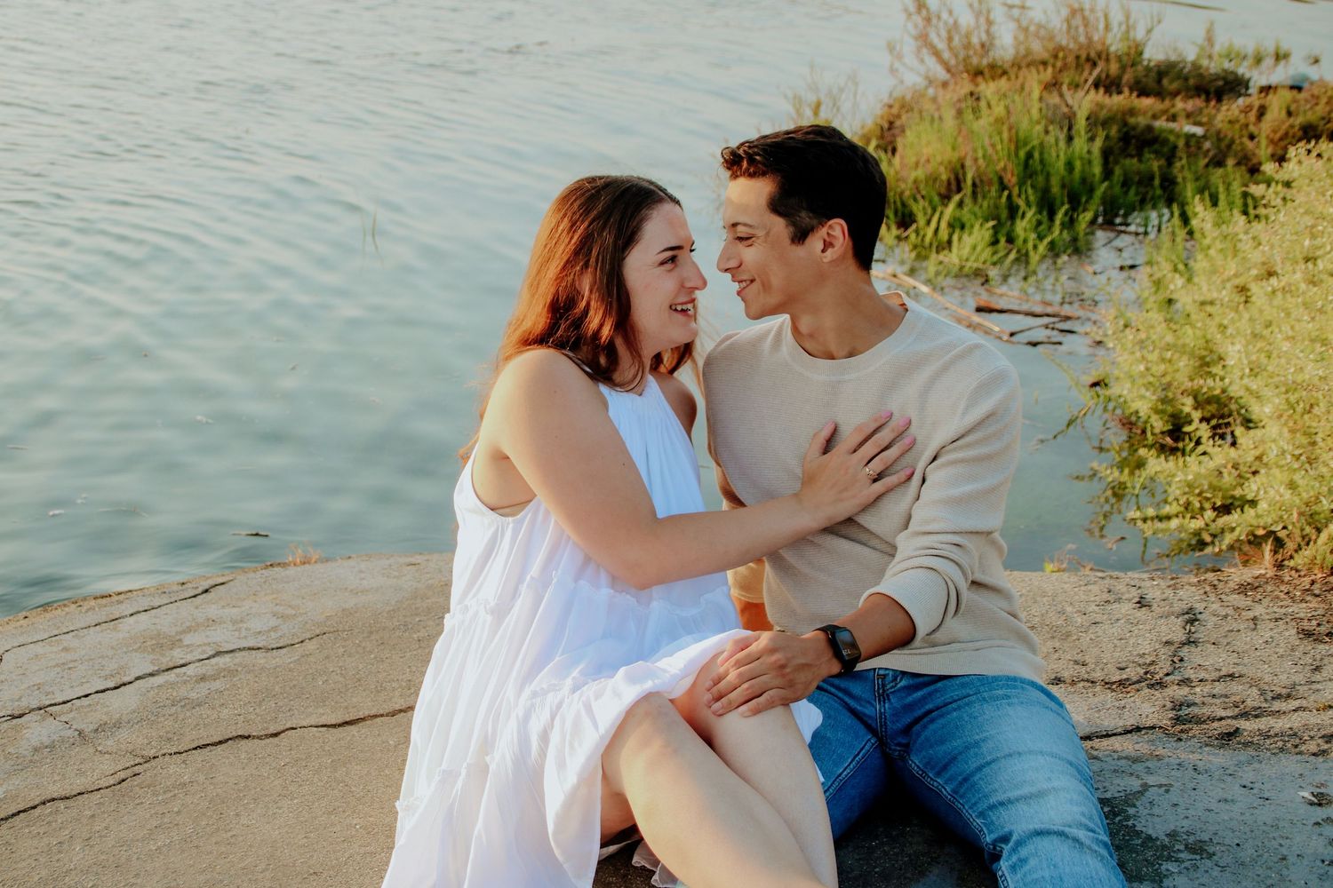Couple in white dress and beige sweater embracing by waterfront during sunset.