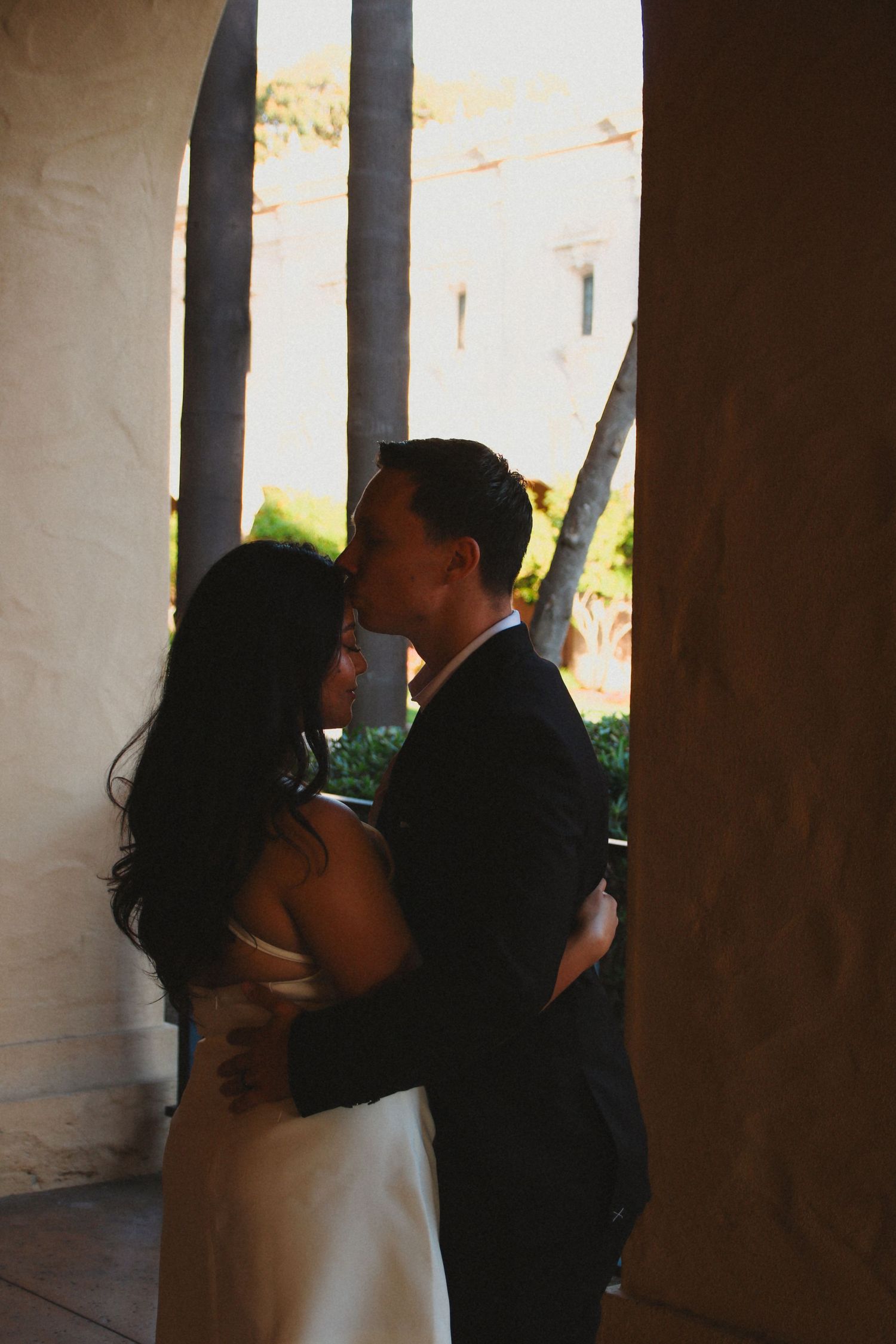 Romantic embrace photographed through stone archway with sunlit background.