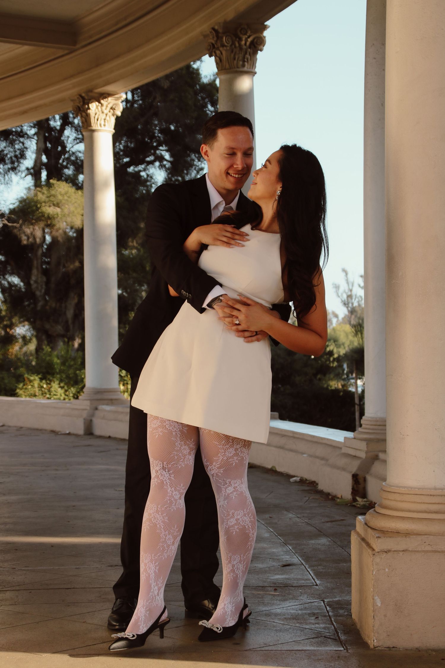 A couple in wedding attire shares intimate moments along a columned patio in warm sunlight.