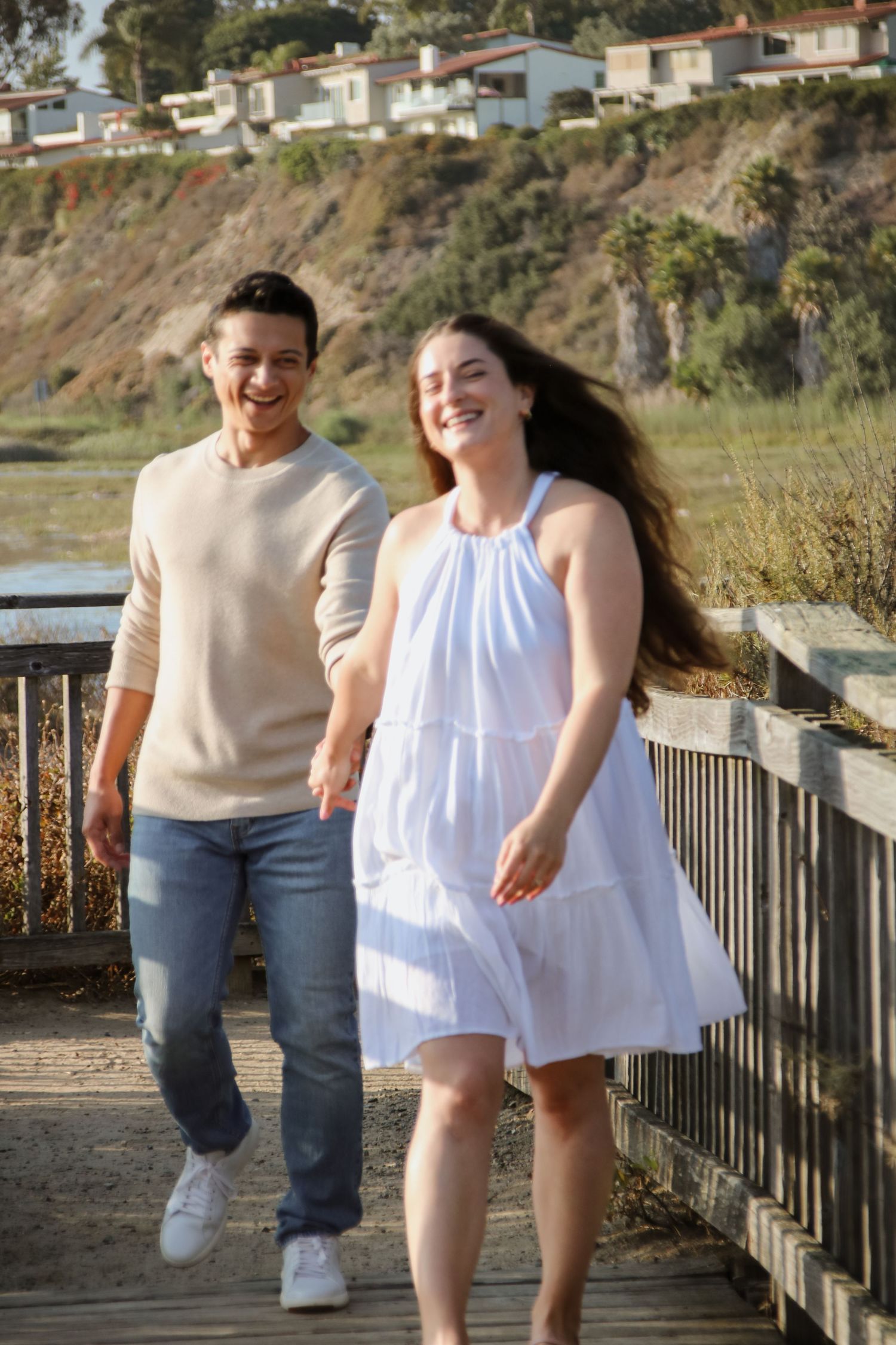 Two people share a joyful moment while walking together on a wooden boardwalk with coastal homes in background.