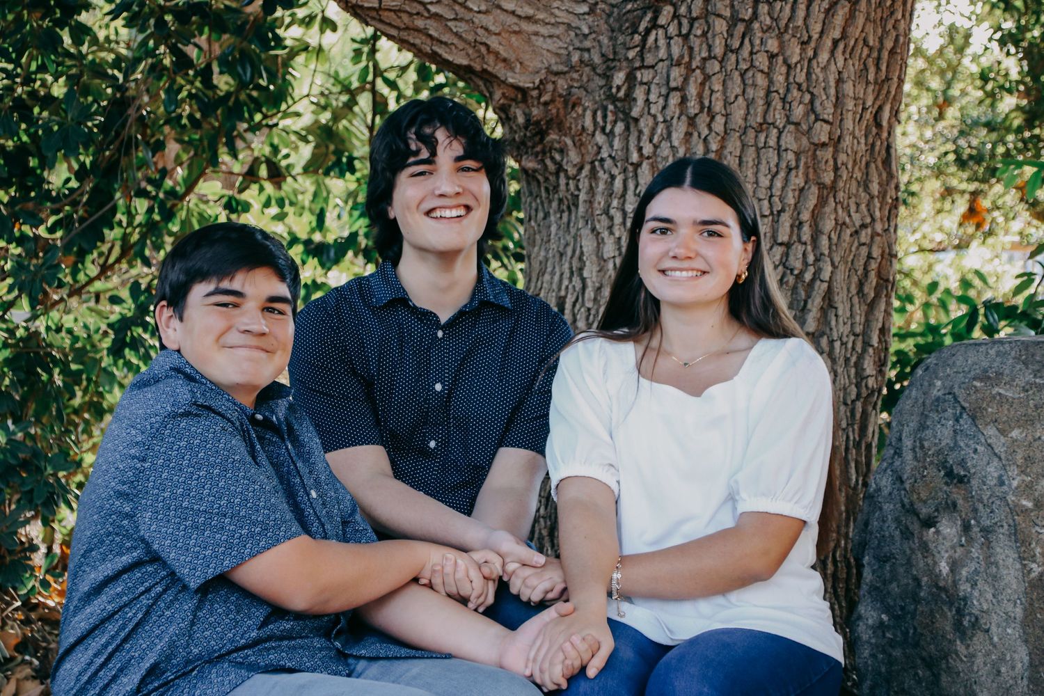 Three teenagers sitting together on rock by tree trunk during outdoor photo session.