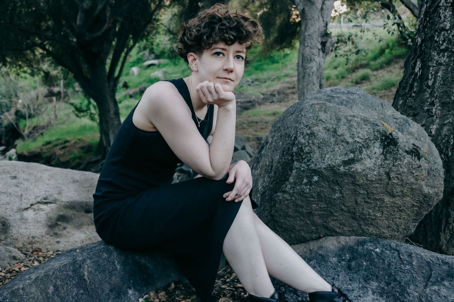 Person in black outfit sitting thoughtfully on large rocks in a natural outdoor setting.