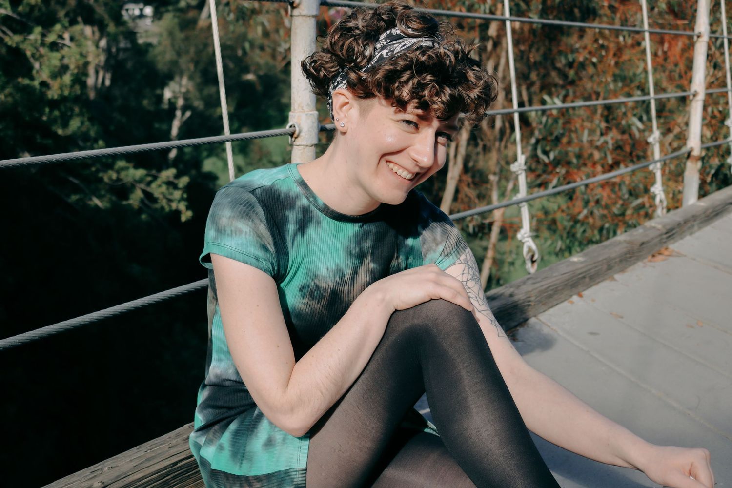 A person laughs while sitting on a wooden bridge deck wearing a green tie-dye outfit.
