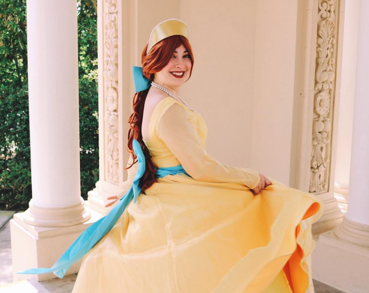 A cosplay of a princess in a flowing yellow ballgown with turquoise sash poses by elegant columns.