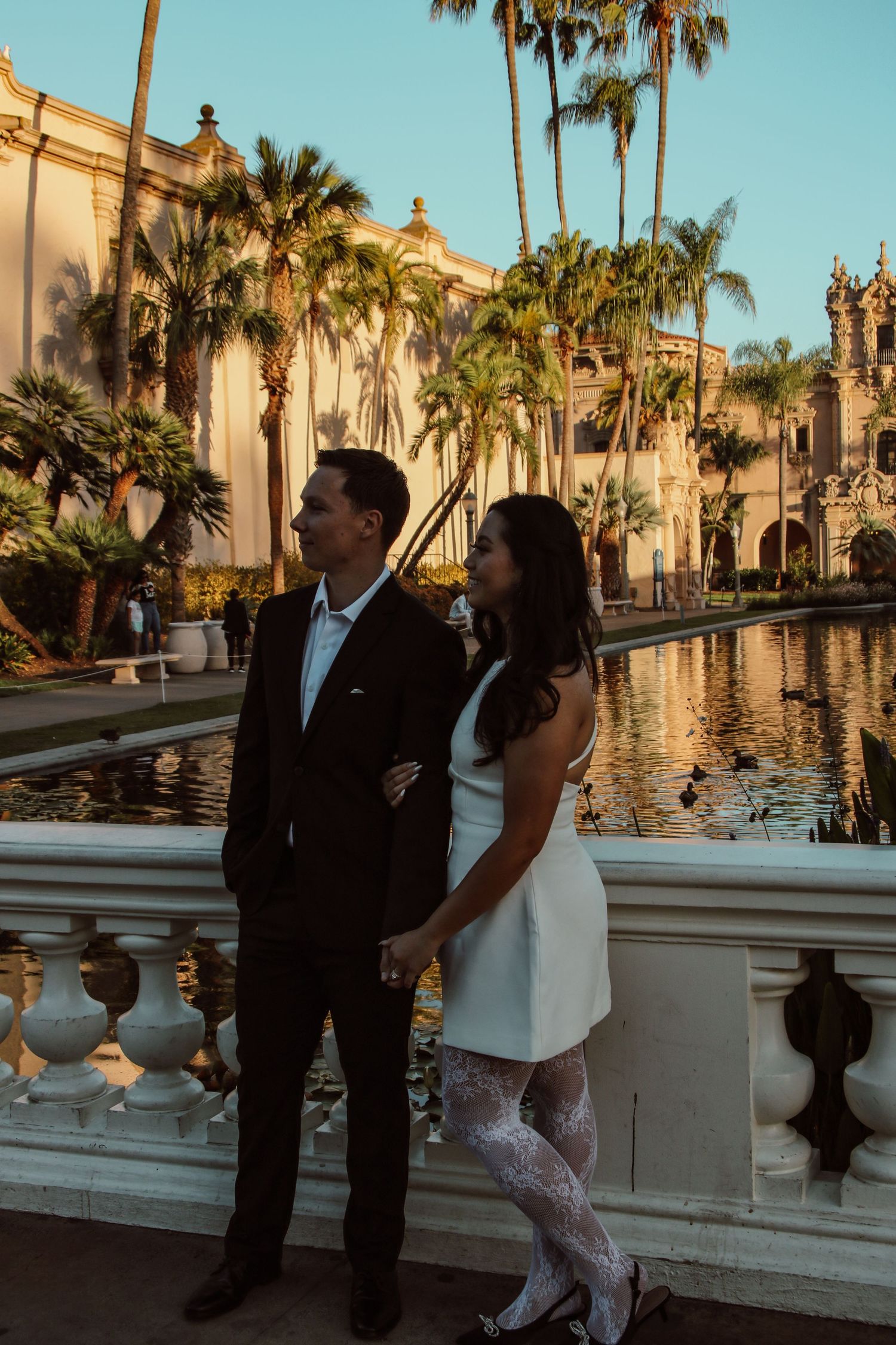 Stylish pair posing by reflecting pool with palm trees and Spanish colonial architecture at sunset.