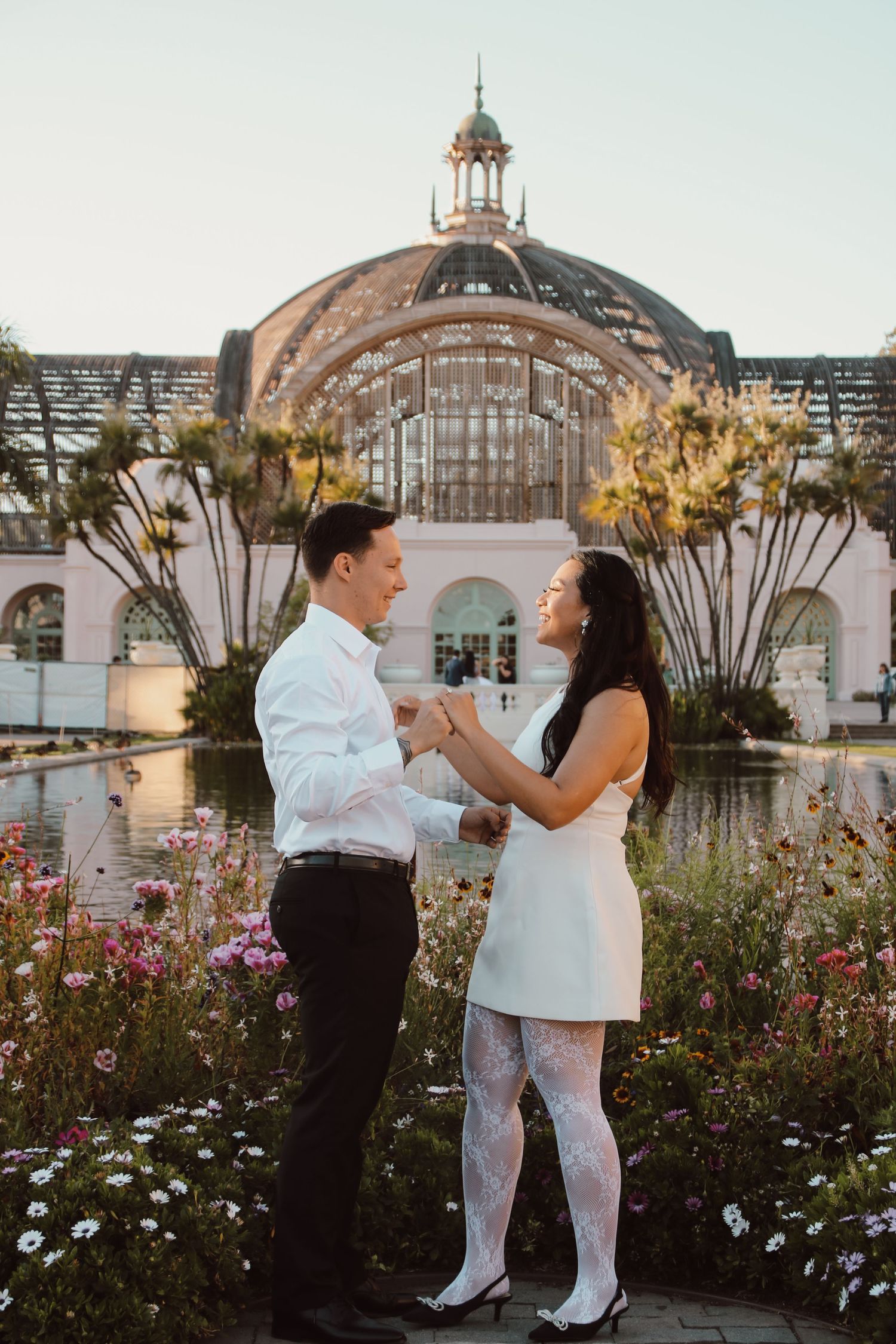 A couple poses romantically in front of a beautiful domed conservatory surrounded by flowers.