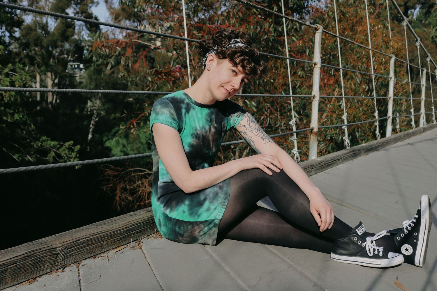 A person in green tie-dye dress and black tights sits casually on a wooden deck.