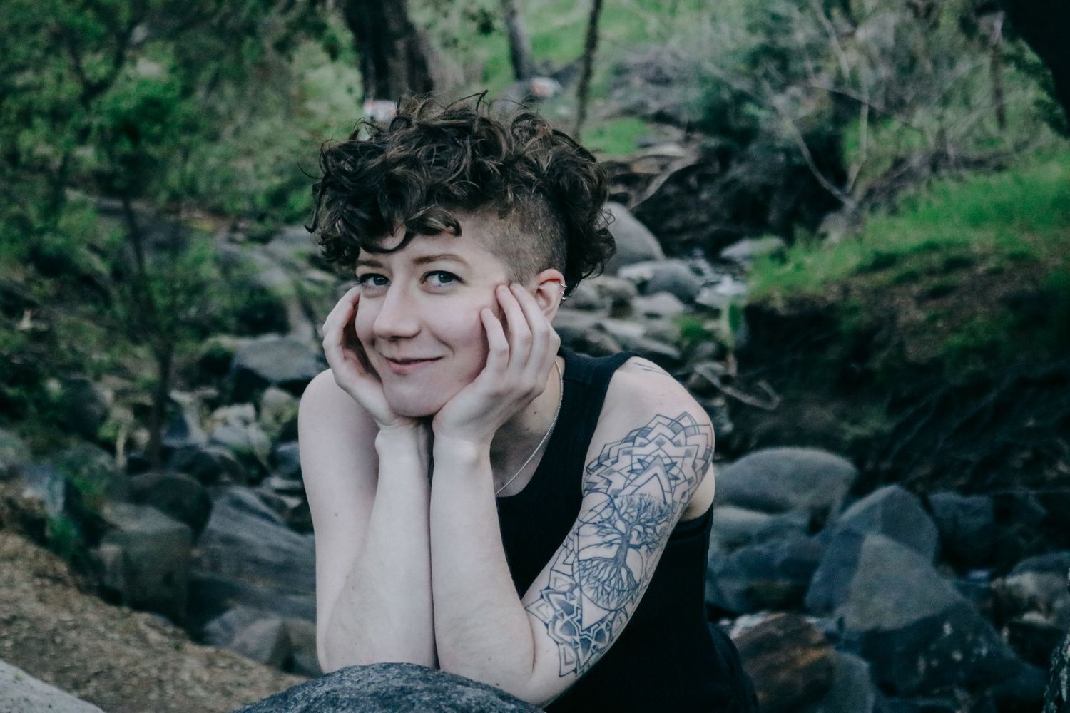 Candid outdoor portrait showing person with short curly hair resting head in hands by rocky stream.