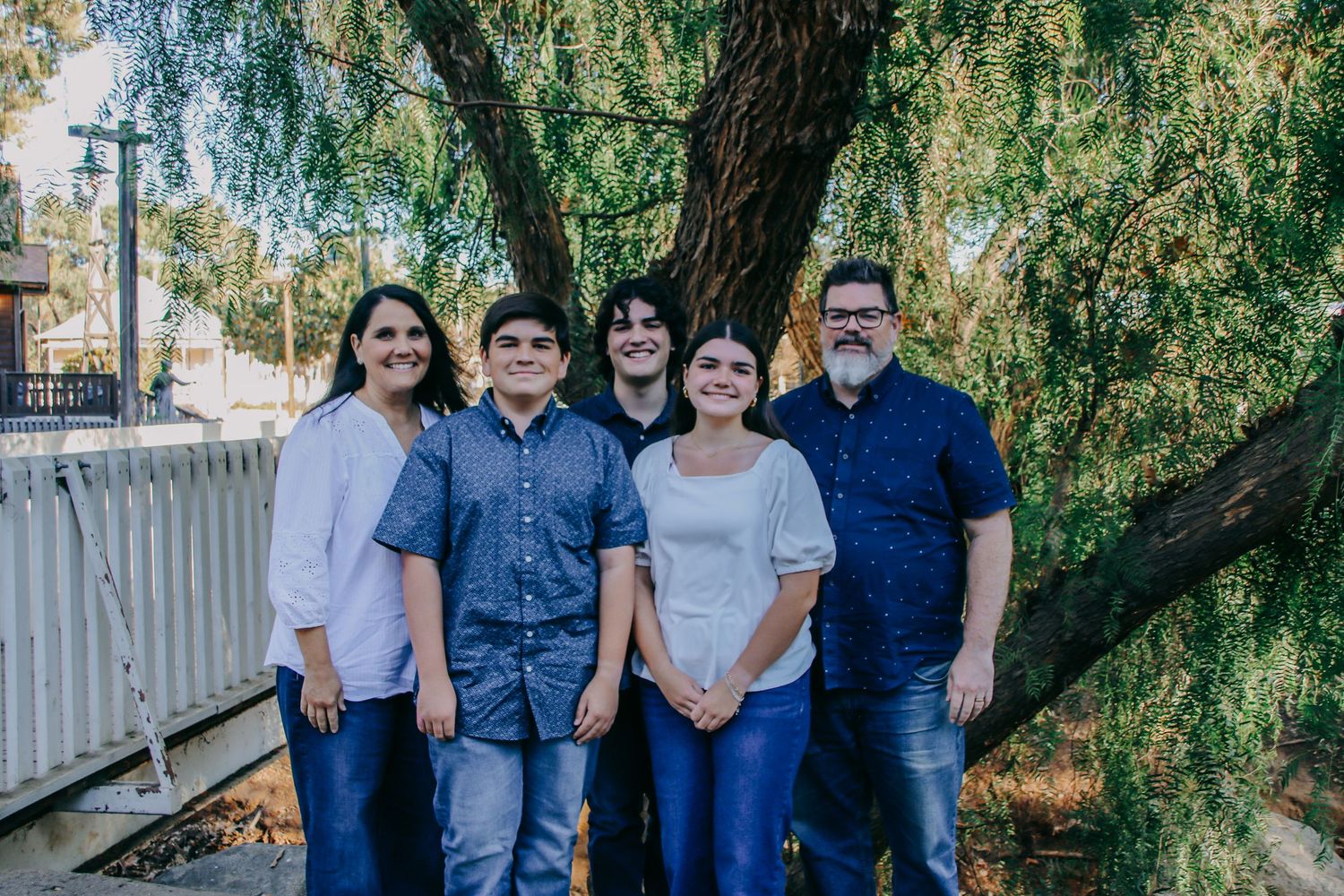 Casual family photo of five members standing together on wooden deck under willow tree.