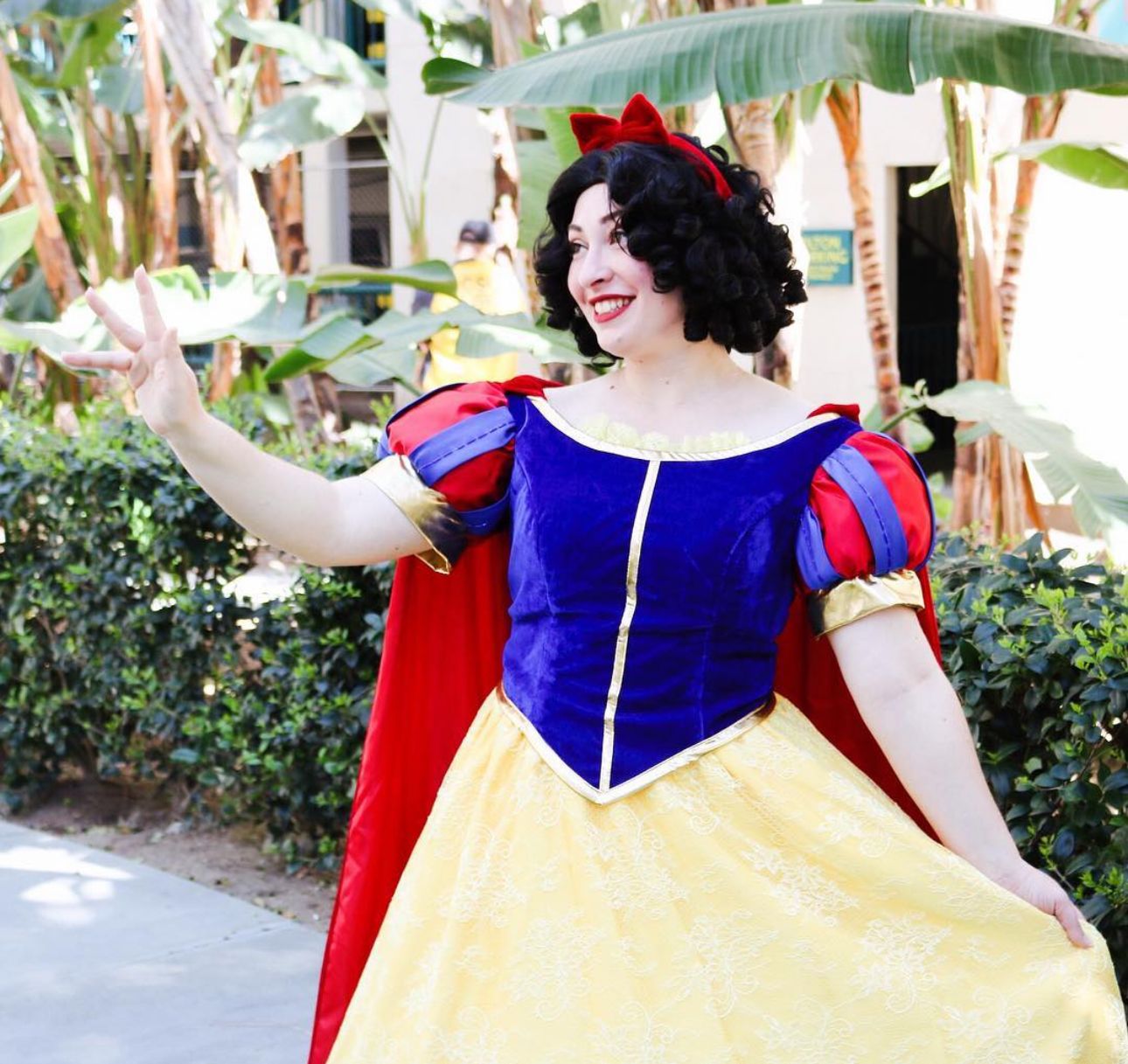 A cosplayer wearing a classic blue and yellow princess dress poses in a garden setting.