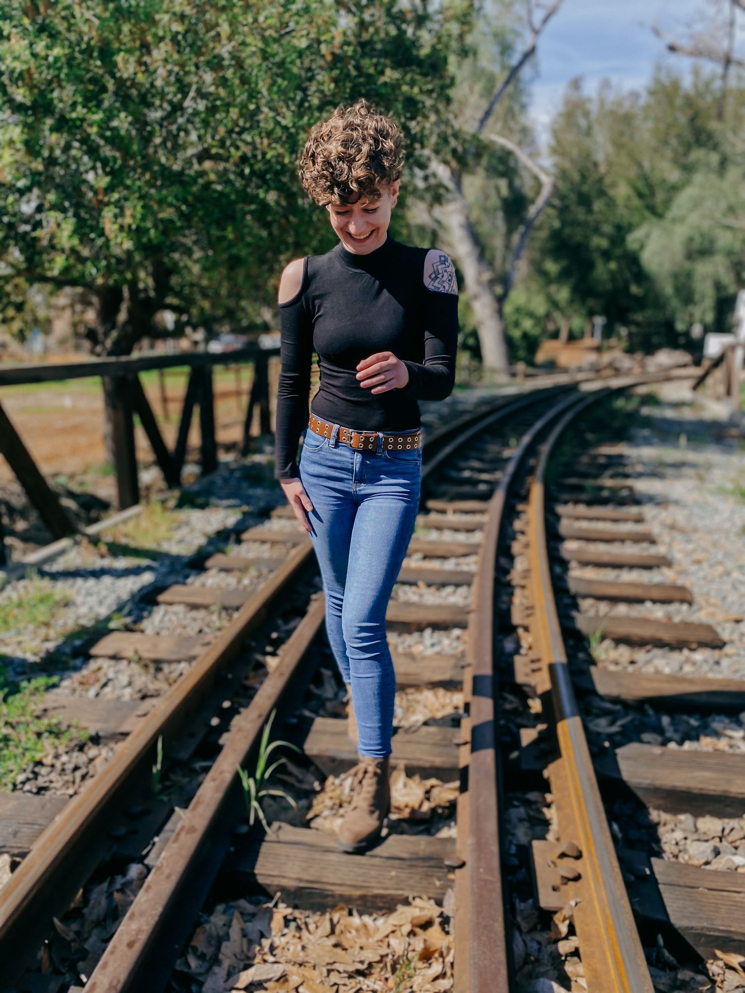 A person in a cold-shoulder black top walks along rustic train tracks surrounded by trees.