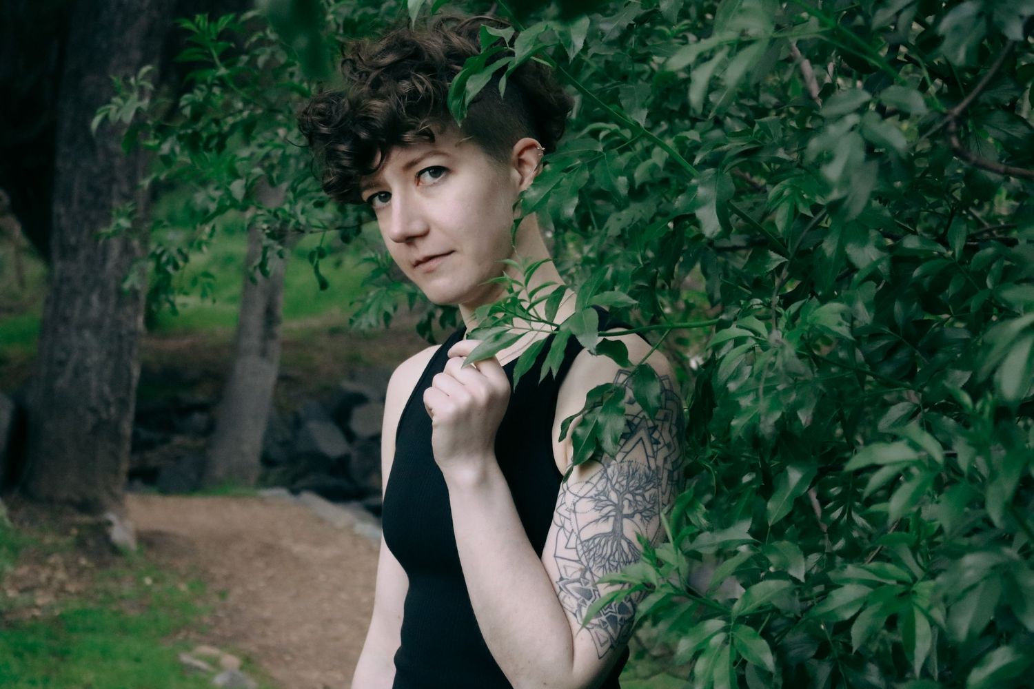 Person in black sleeveless top poses among green foliage showing intricate arm tattoo.