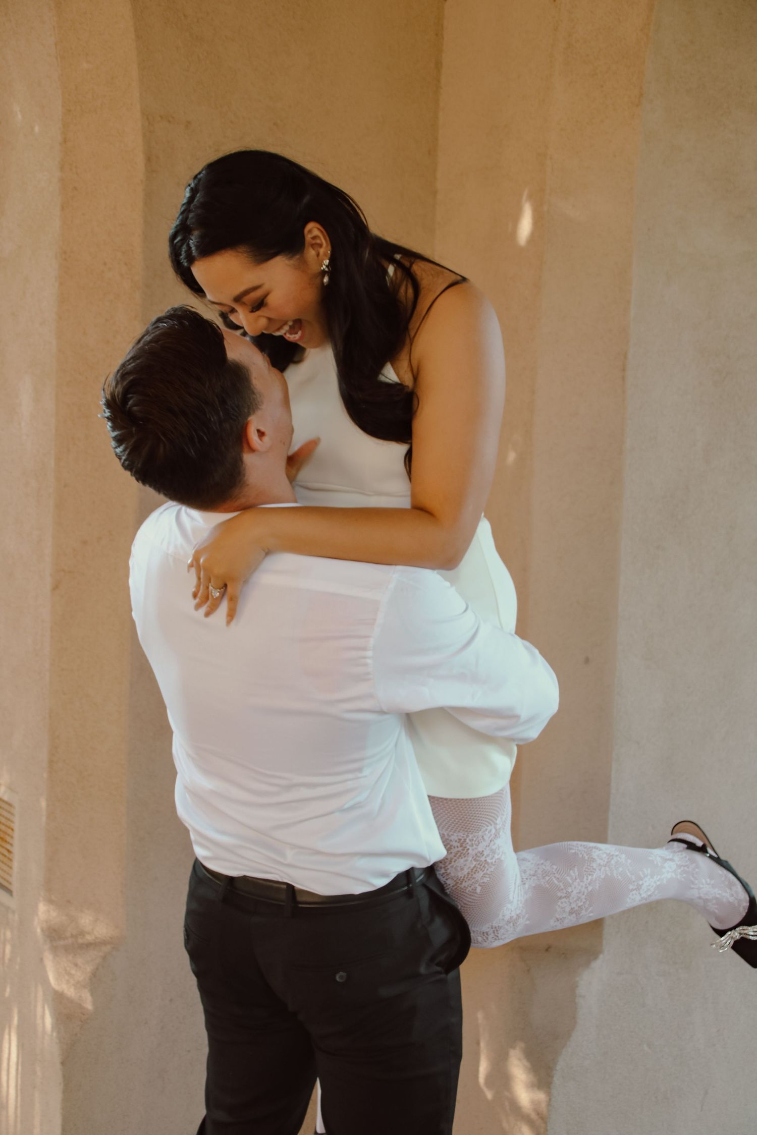 Intimate portrait of couple embracing in warm sunlight against neutral wall.