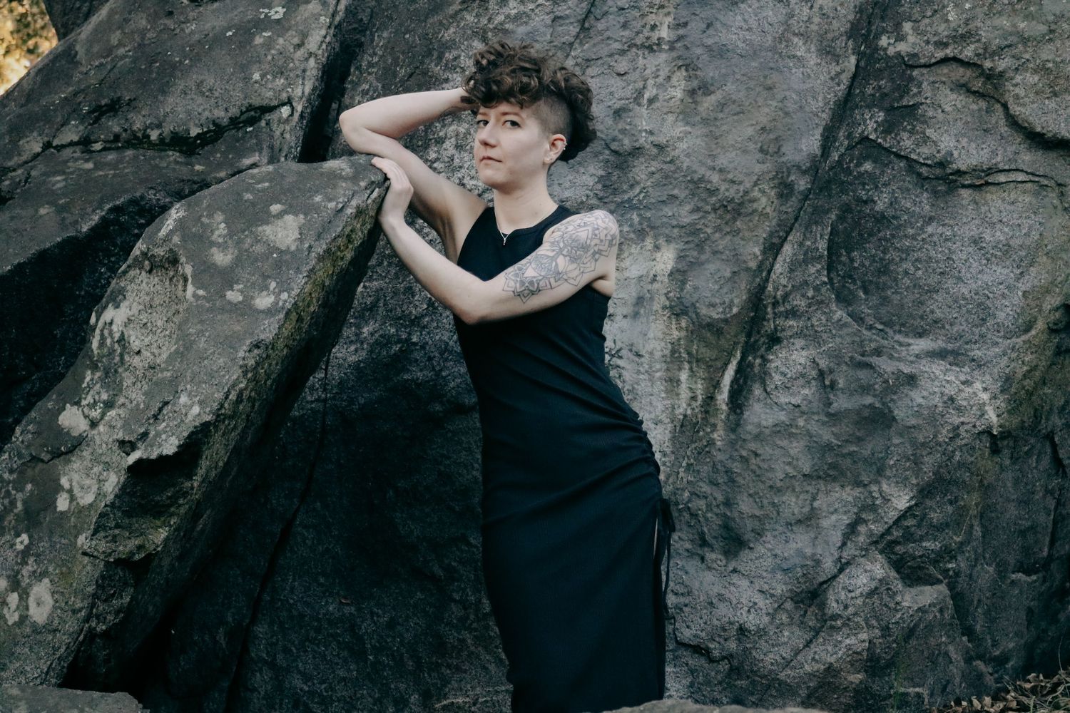 Person in dark outfit poses expressively against textured stone background.