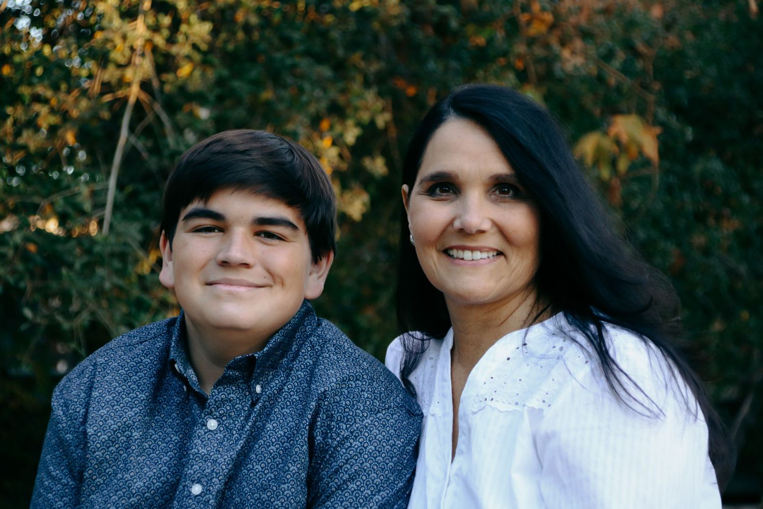 Close-up portraits of two people sitting together outdoors with golden autumn foliage behind them.