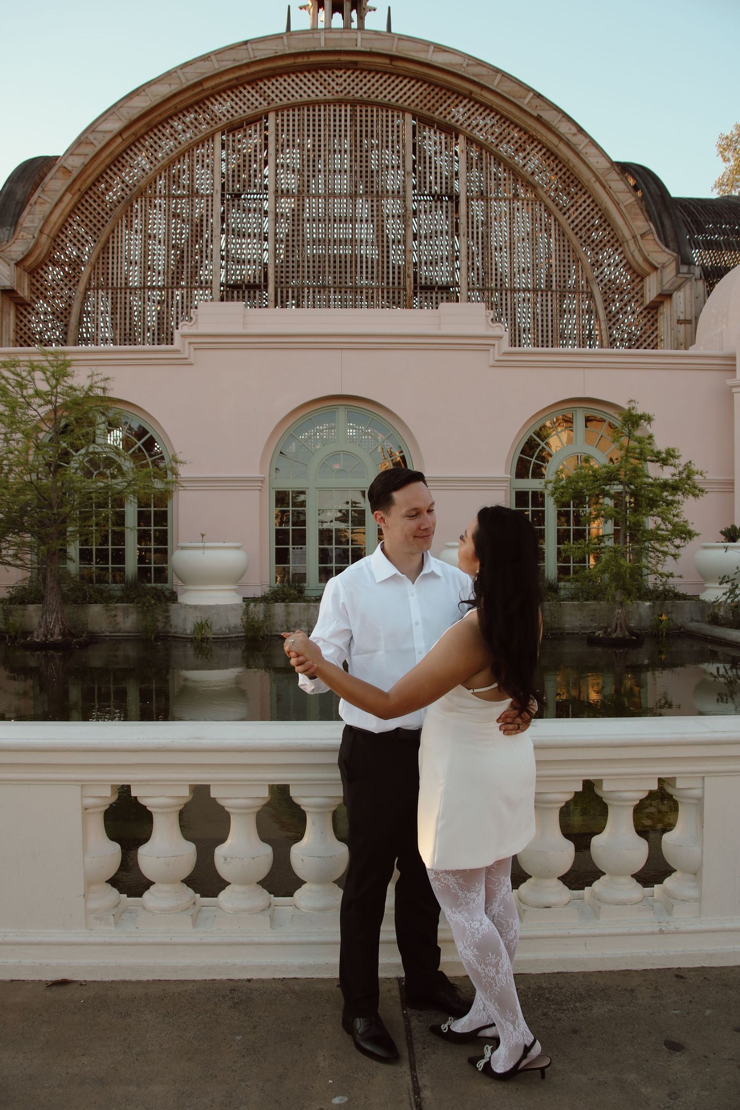 Couple dancing in formal attire by an ornate architectural structure with arched details and reflecting pool.
