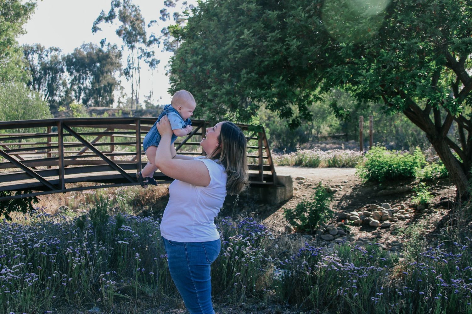 A parent lifts a baby against a rustic bridge backdrop with wildflowers in bloom.