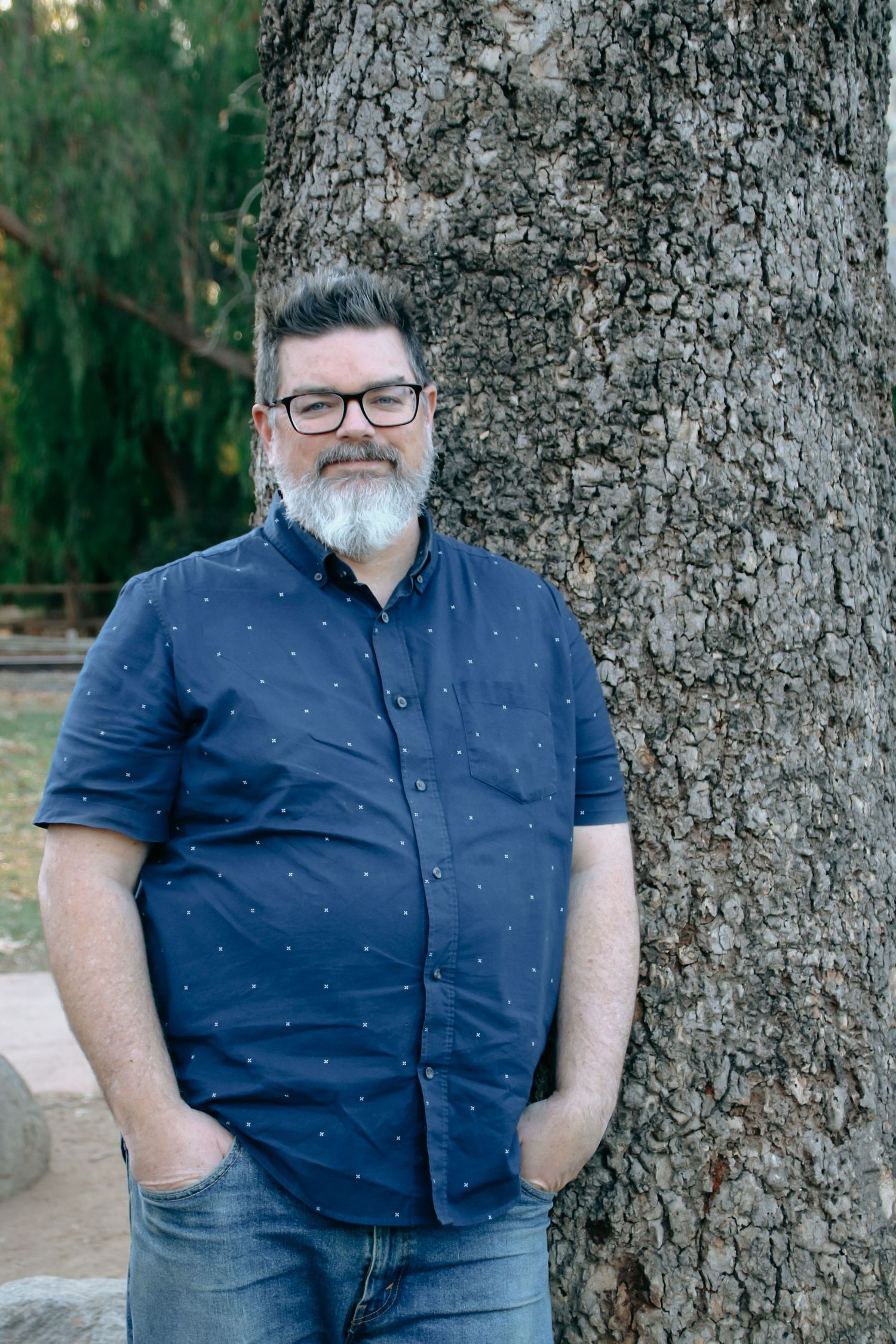 A person with beard and glasses poses casually against a tree in an outdoor setting.