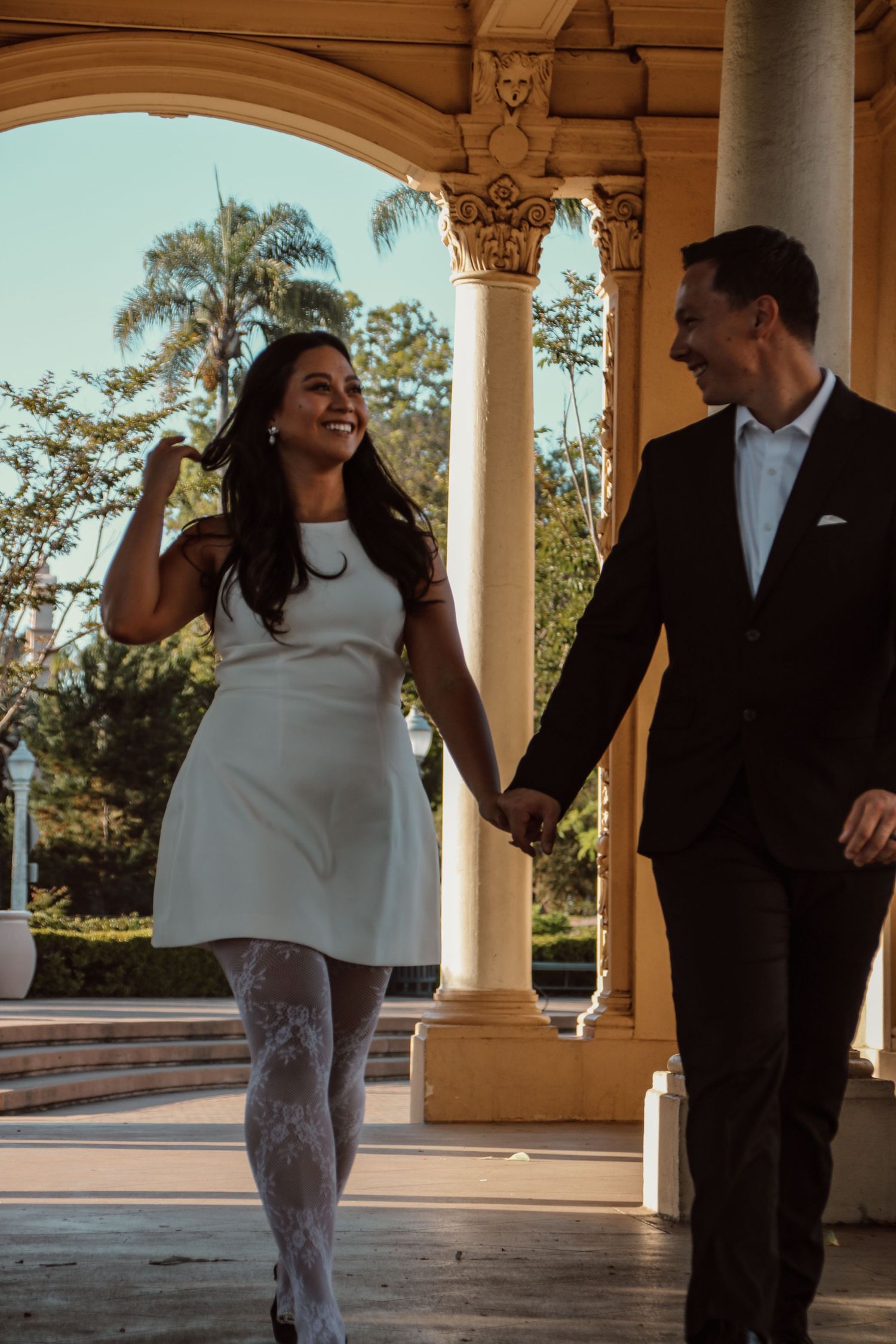A couple enjoys a playful moment while walking through a sunlit colonnade.