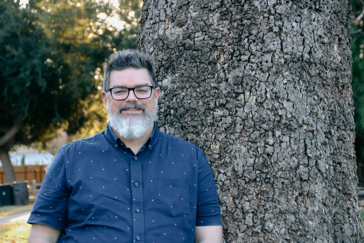 A man with gray beard and glasses stands next to a textured tree trunk in casual clothing.