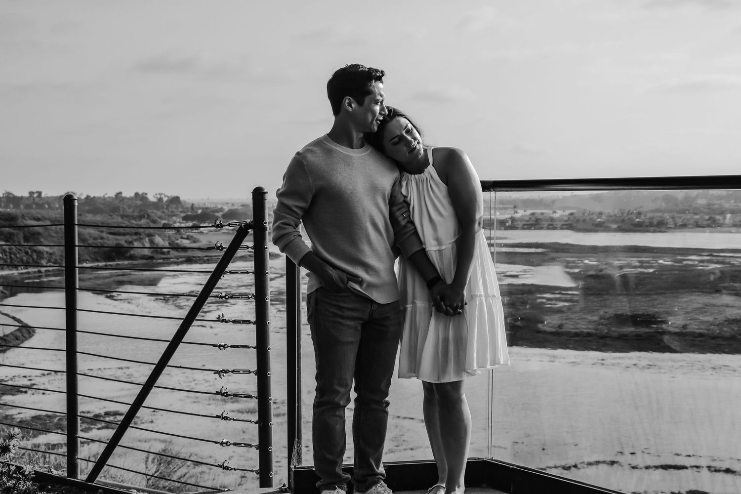 An intimate black and white portrait of a couple embracing on a coastal viewing platform.