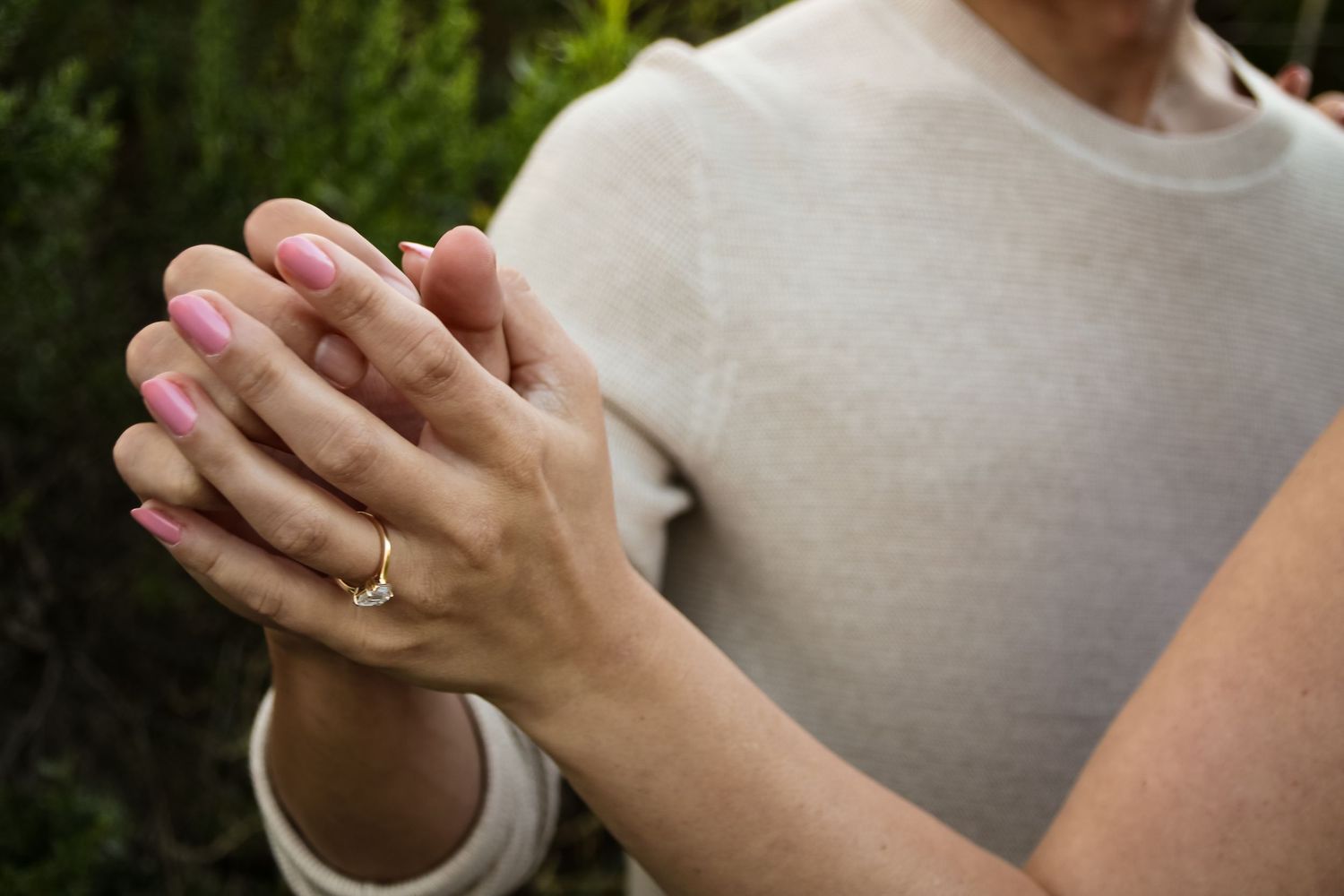 Close-up of clasped hands with an engagement ring visible against a beige sweater background.