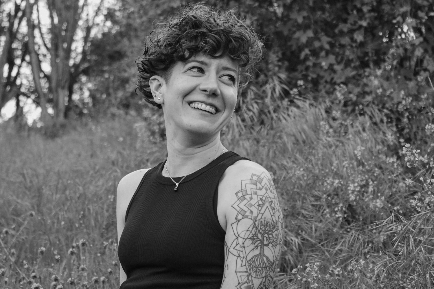 Black and white portrait of person in sleeveless top with visible arm tattoo against natural background.