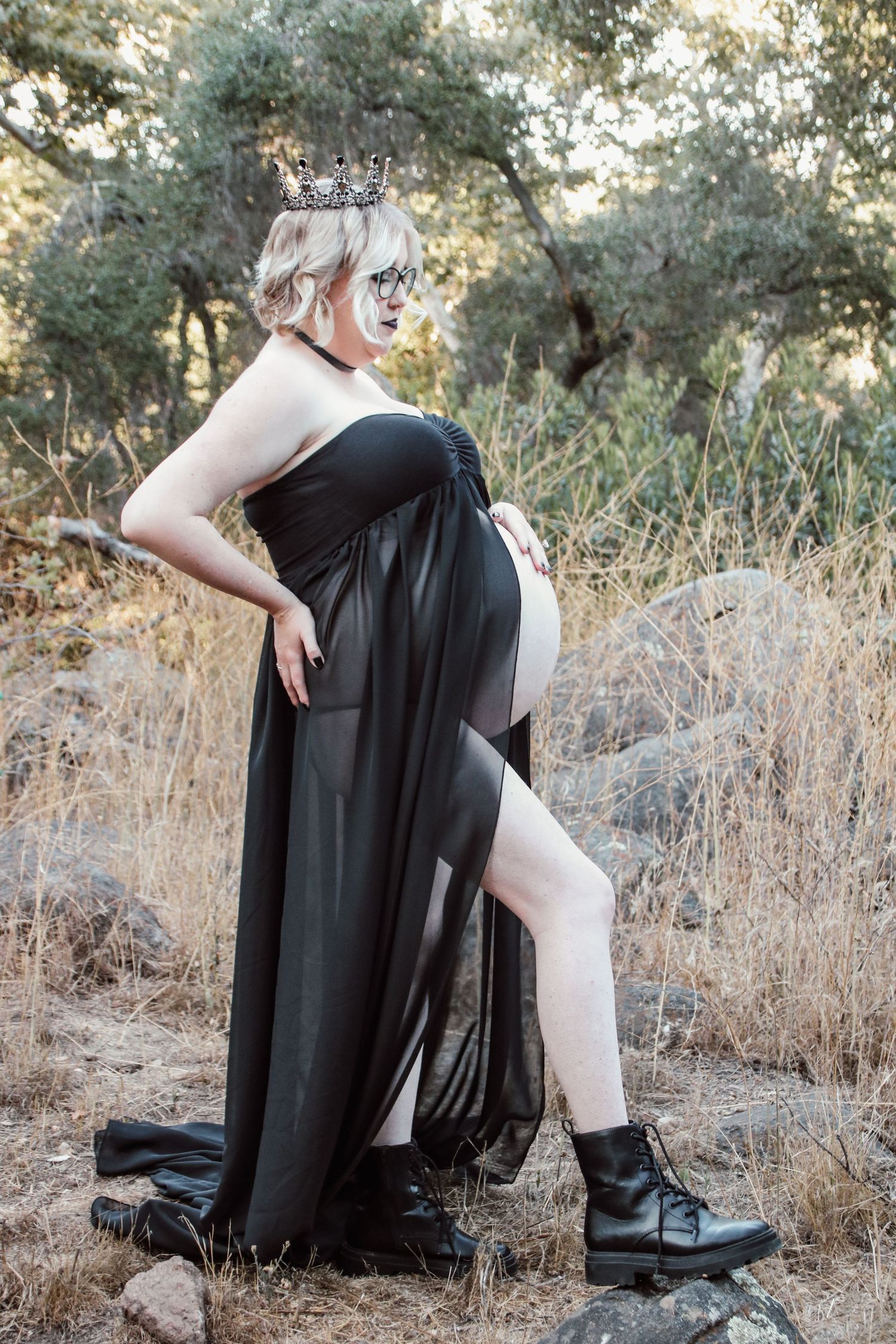 A maternity portrait showcases a flowing black dress and combat boots in a rustic outdoor setting.