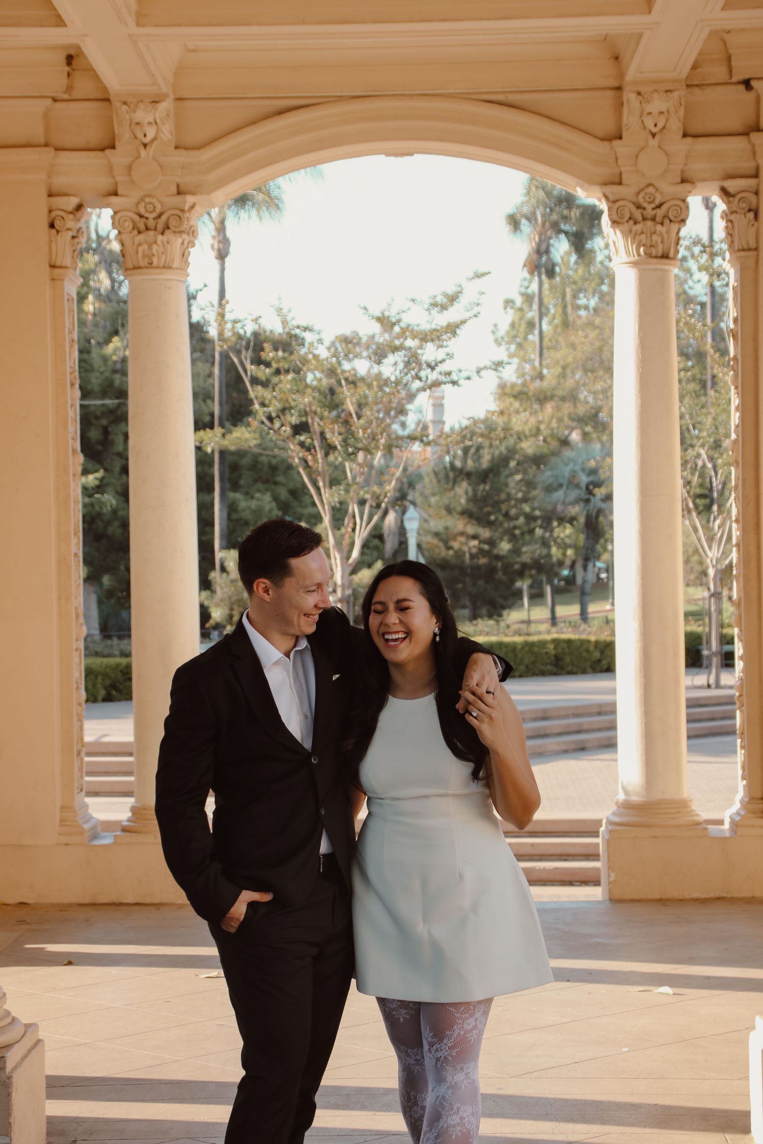 Two people laugh together while posing in an outdoor colonnade with classical architecture.