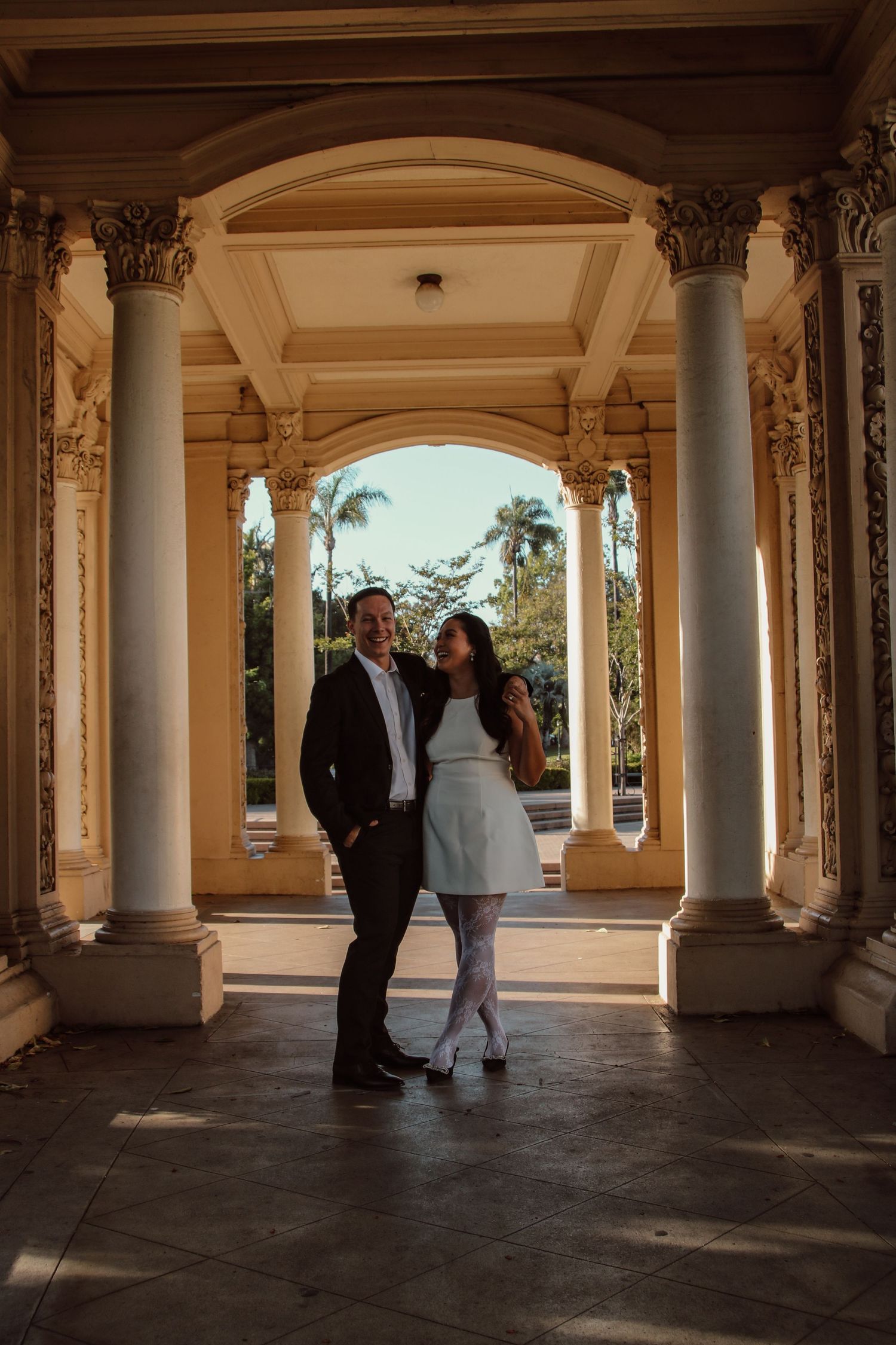 Two people share a romantic moment beneath decorative archways and columns in warm lighting.