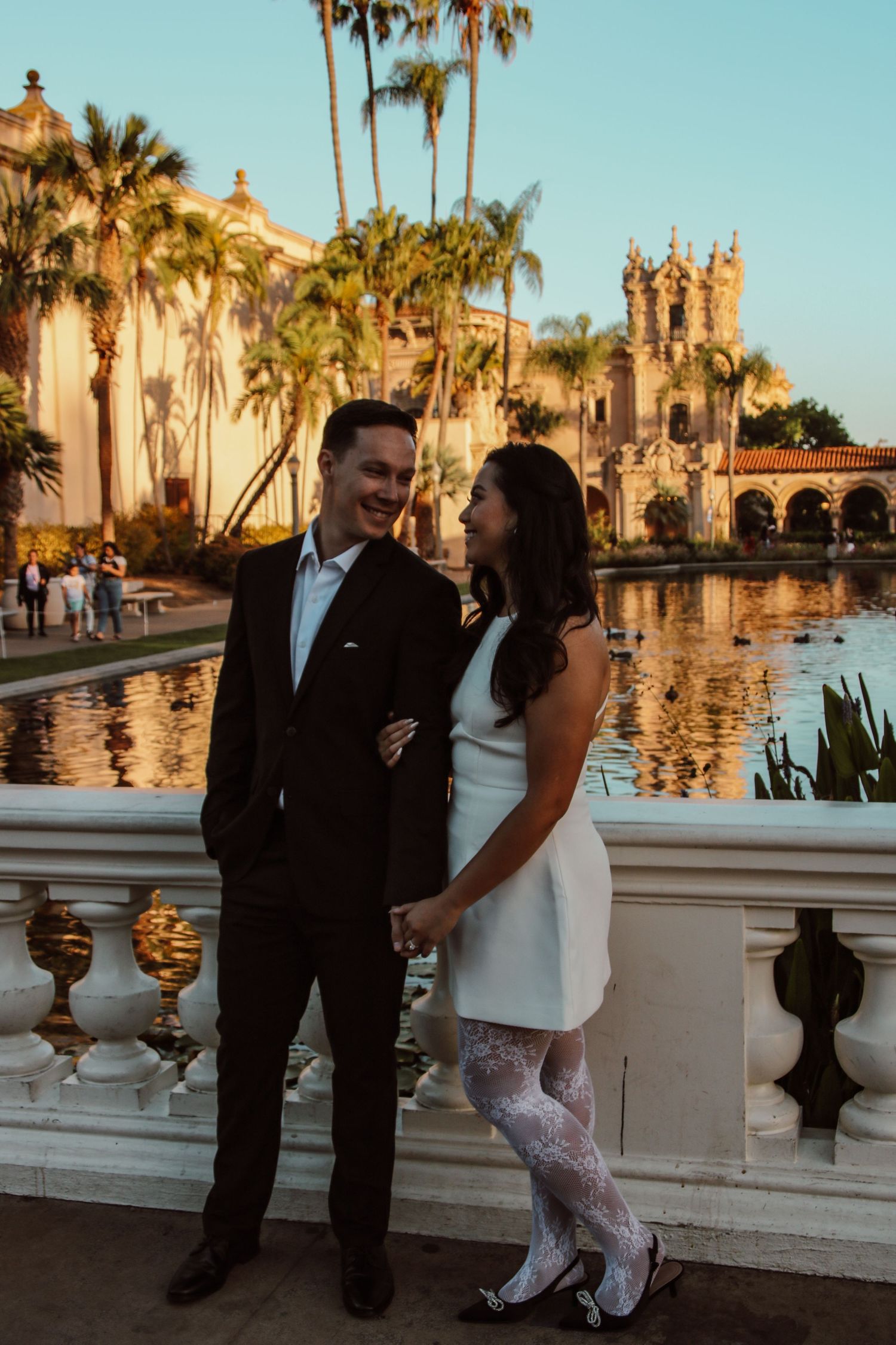 Couple sharing romantic moment by reflective pool with palm trees and Spanish architecture at dusk.