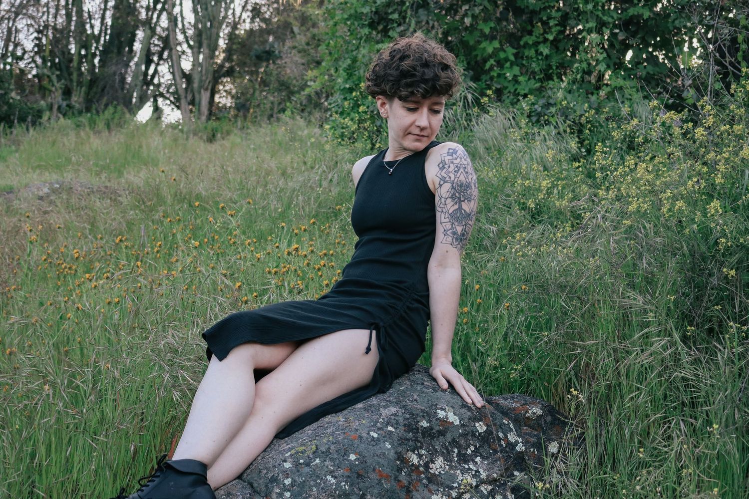 Person in black dress sits on rock against green foliage background.
