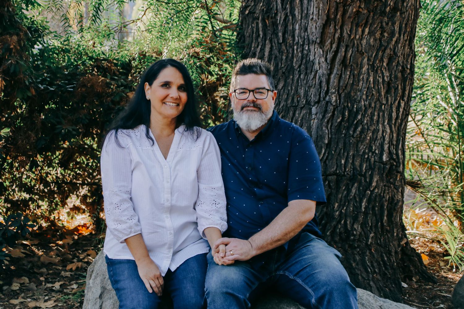 A couple sitting together on a bench against a tree trunk in a sunny outdoor setting.