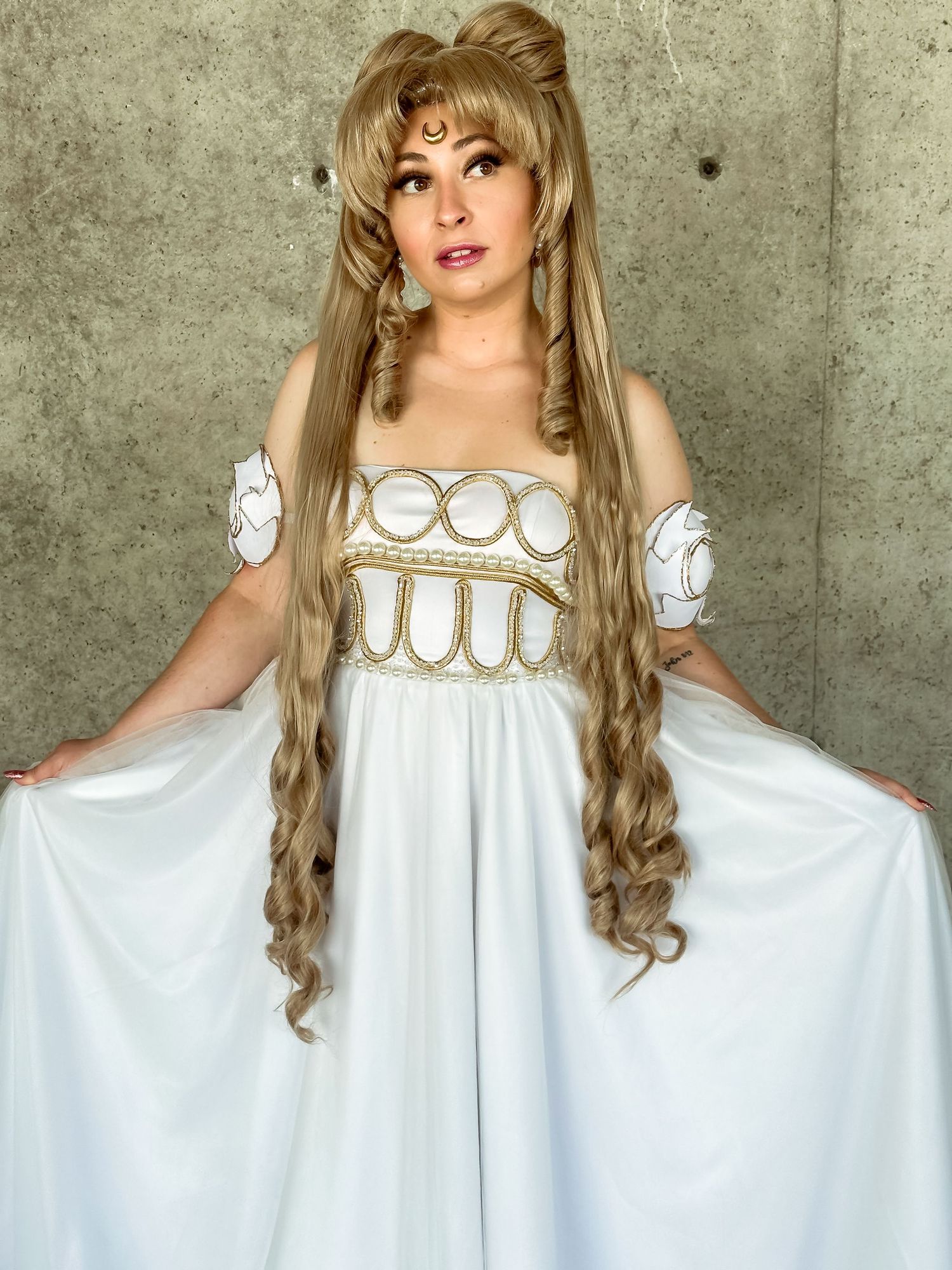 Model in flowing white renaissance-inspired dress with gold details and long braided hair poses against concrete wall.