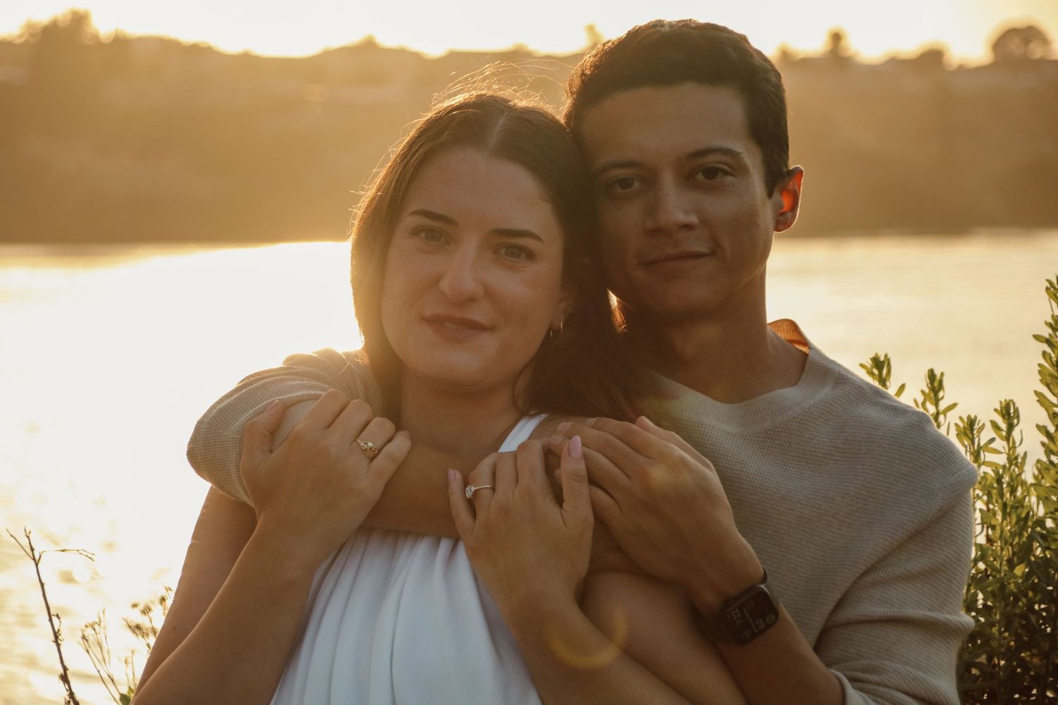 Couple embraces lovingly at golden hour with water and trees in background.