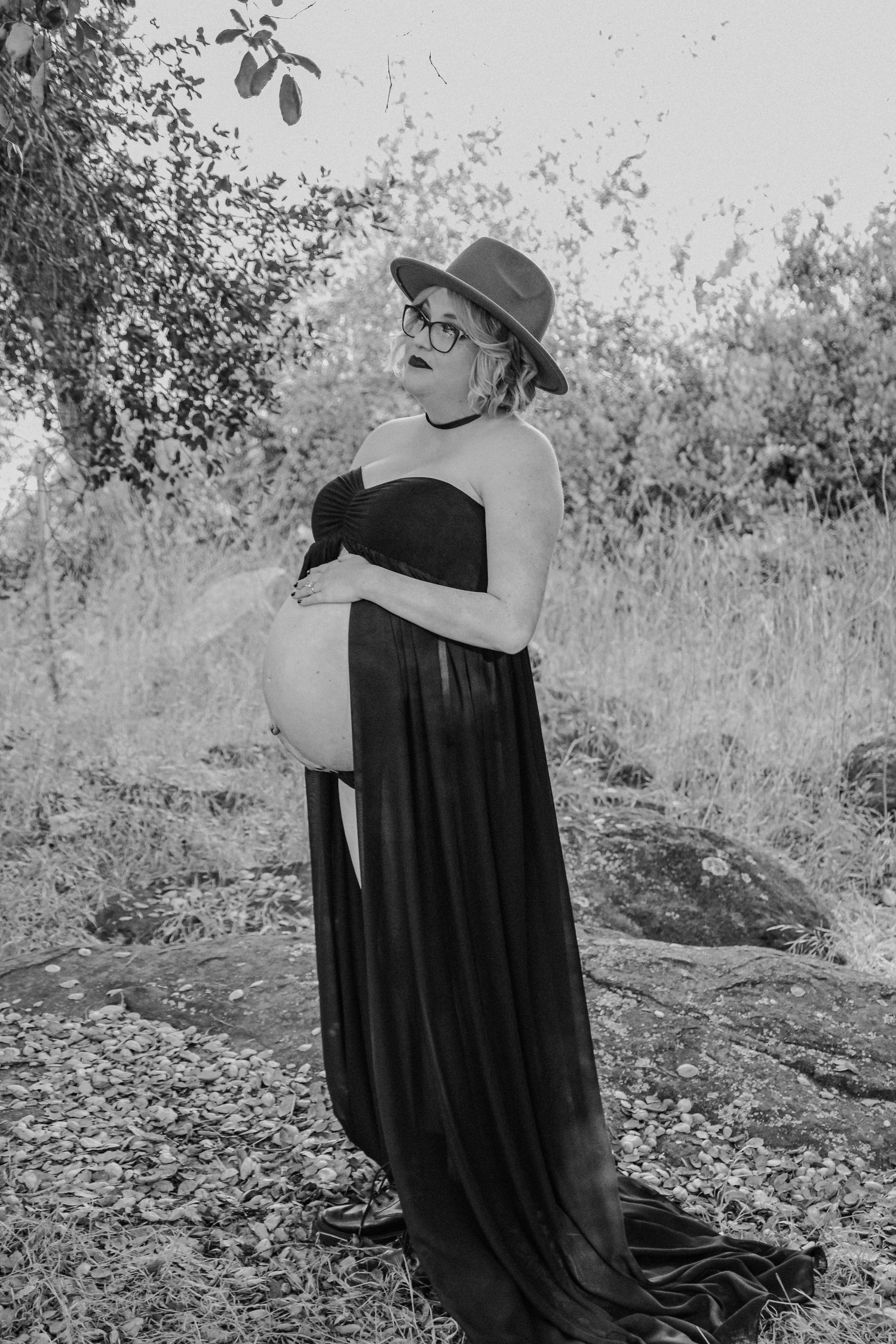 Black and white photo of someone in a dark dress and hat sitting alone on natural rocks in a wooded setting.