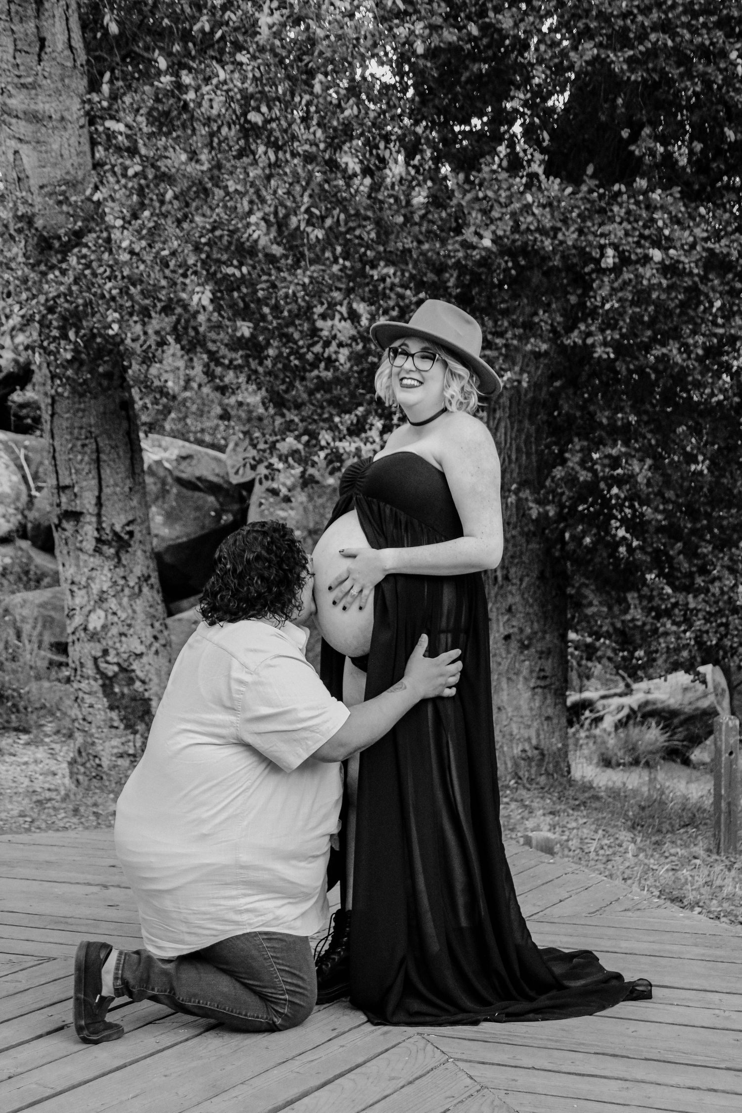 A tender black and white maternity moment captured on a wooden deck surrounded by trees.