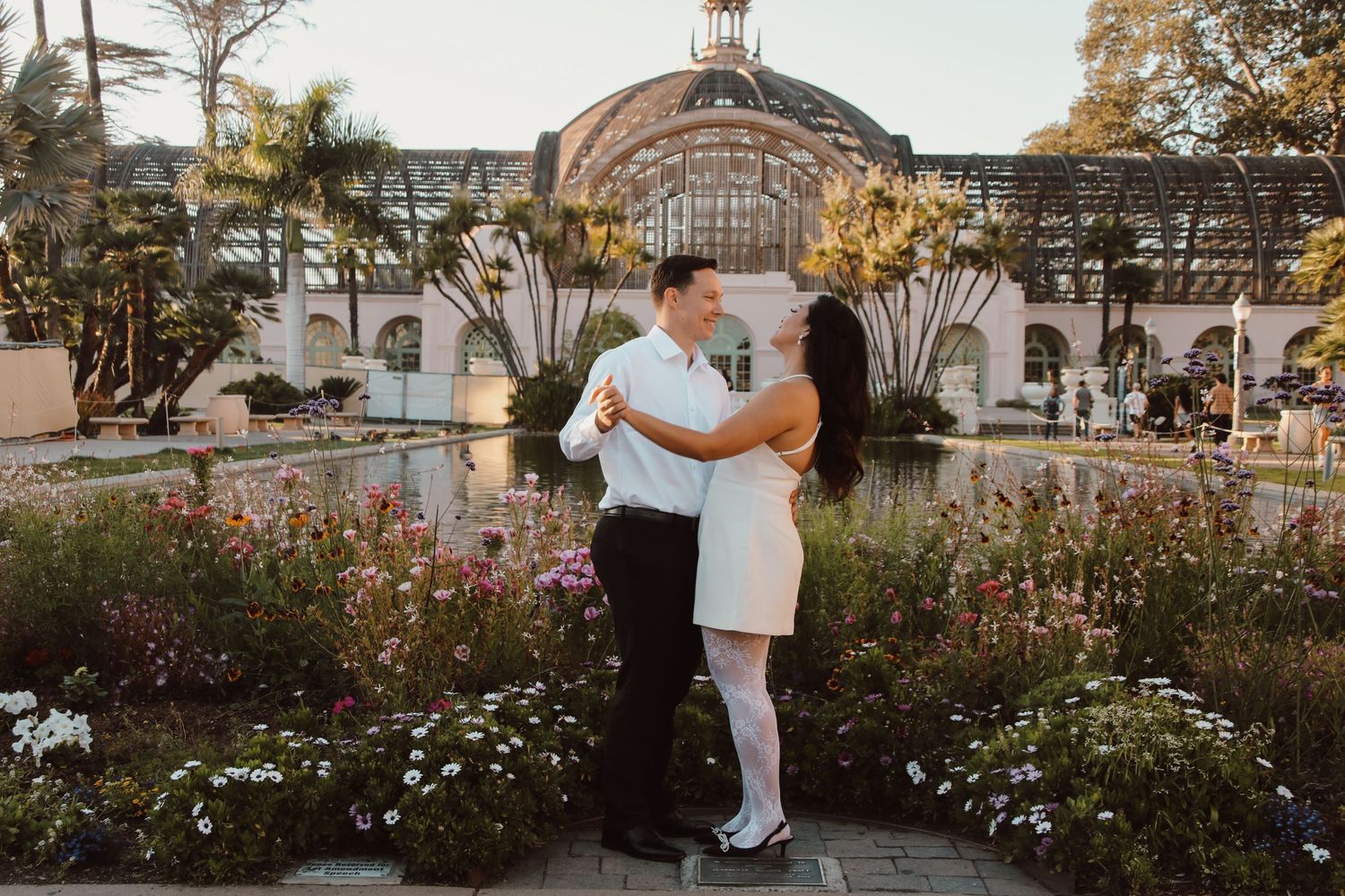 A romantic embrace in front of an elegant glass conservatory surrounded by blooming flowers at sunset.