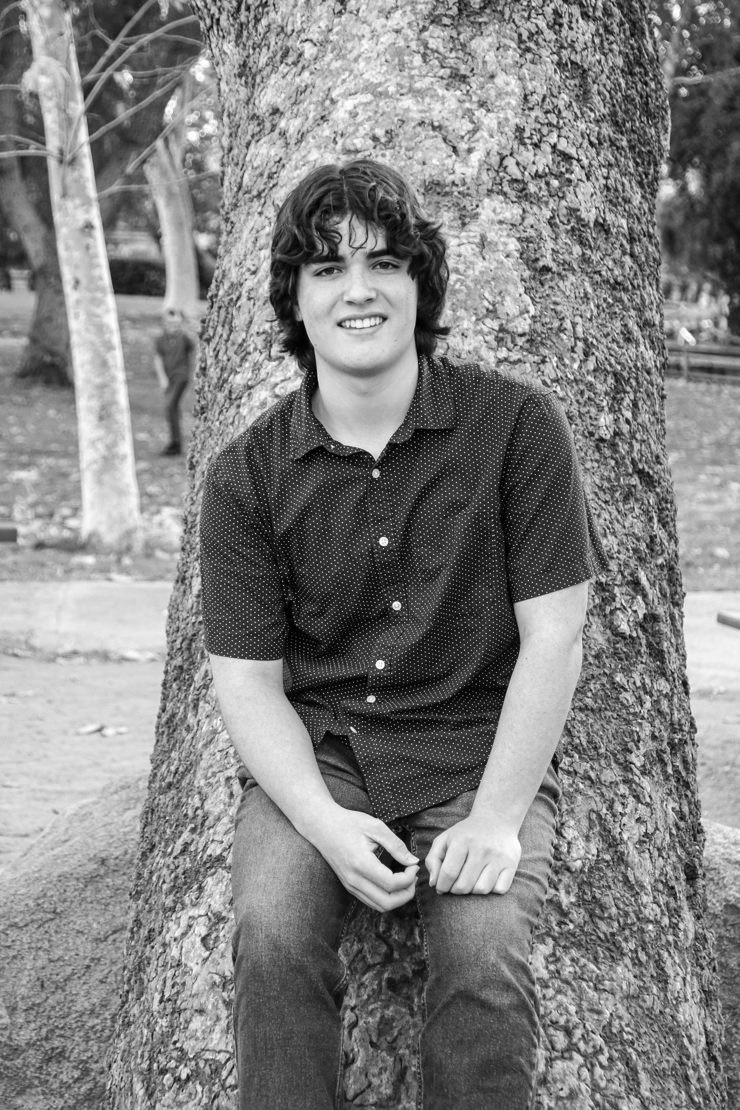 A casual portrait of someone sitting by a tree trunk wearing jeans and a dark button-down shirt.