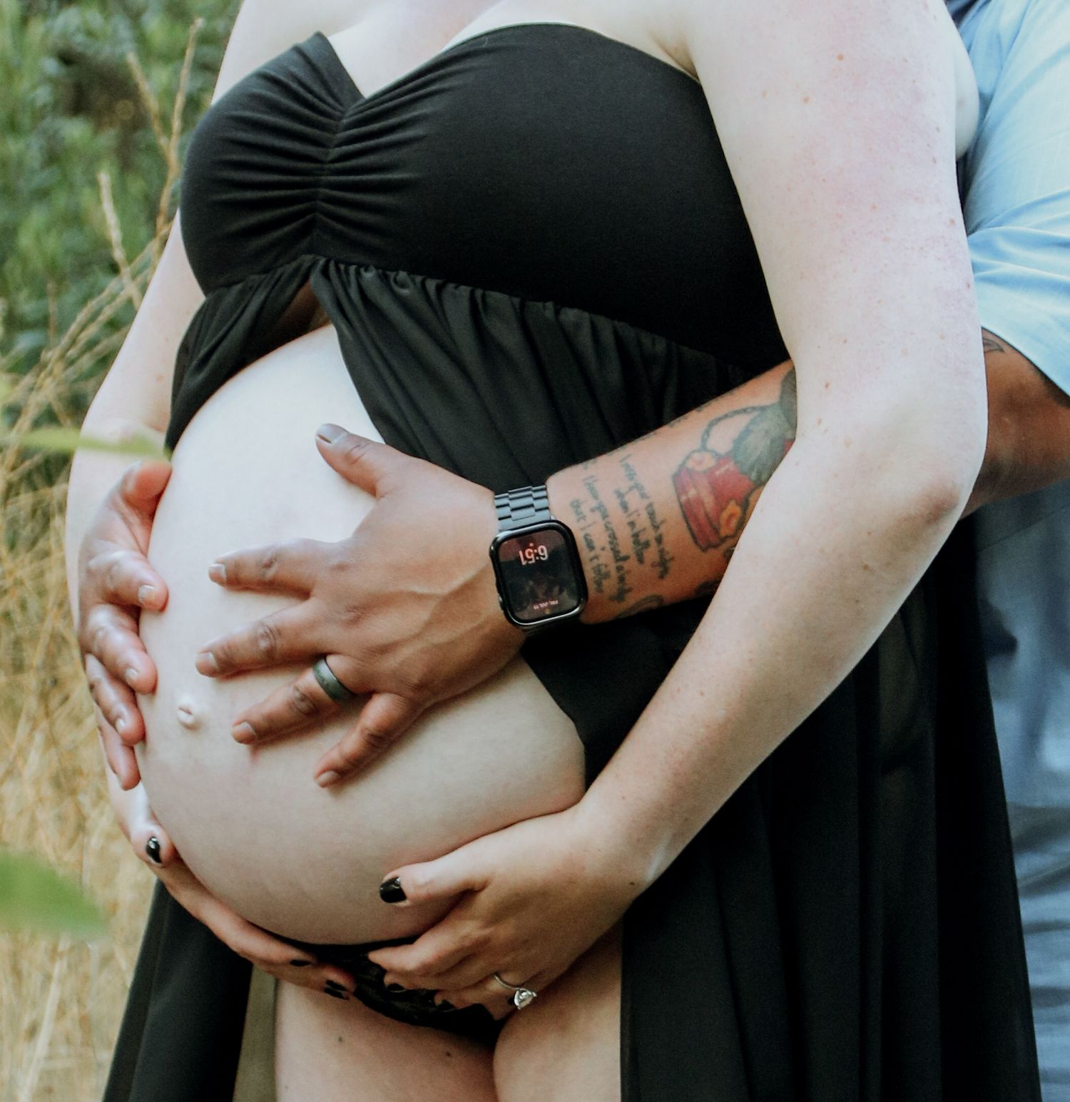Pregnant woman in black dress with Apple Watch holding her belly with hands in outdoor maternity photo.