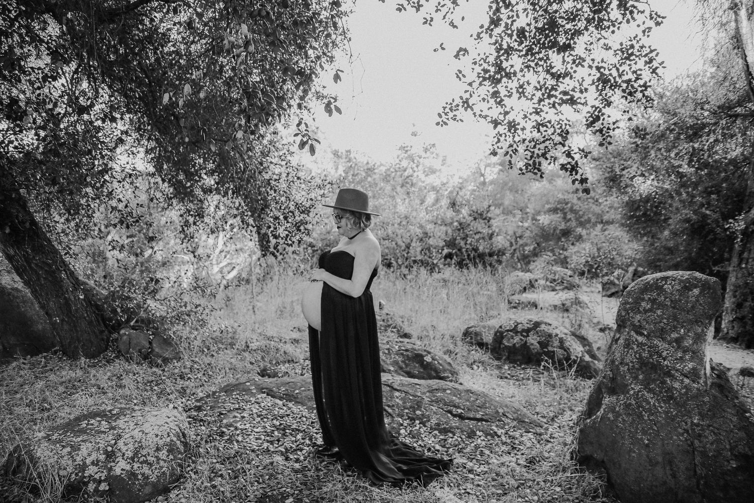 A figure in a long black dress stands in a romantic naturalistic garden setting captured in black and white.
