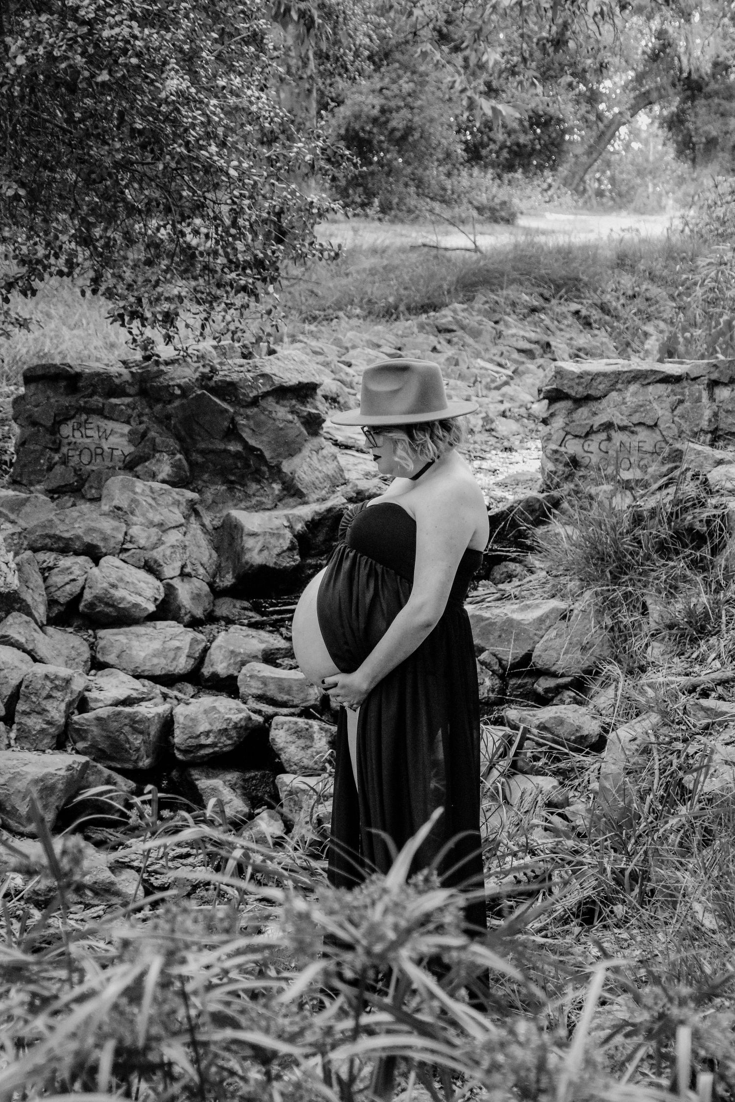 A black and white maternity photo featuring a pregnant woman in a dress standing near a stone wall.