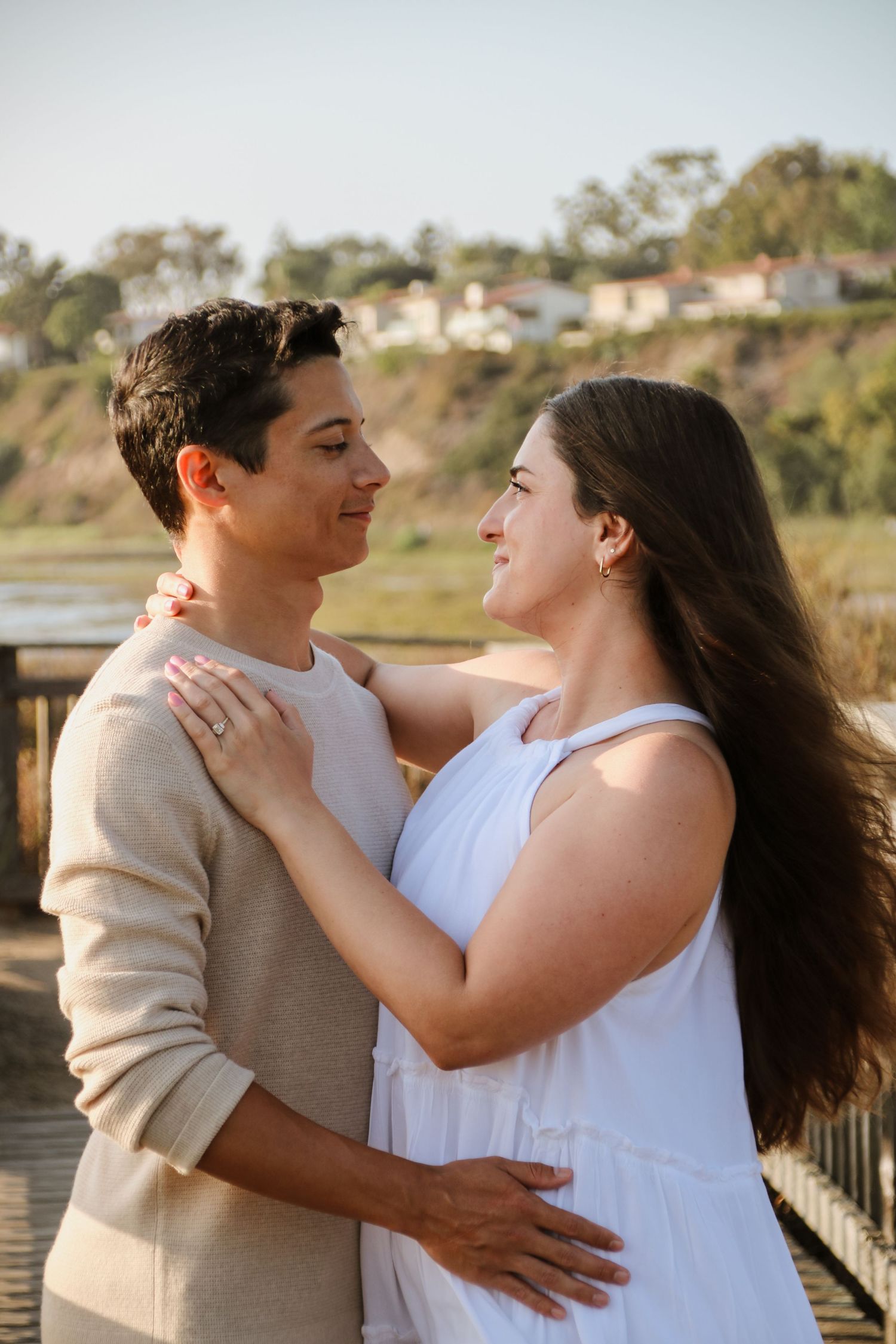 A couple embraces lovingly on a boardwalk during golden hour with houses visible on the hillside.
