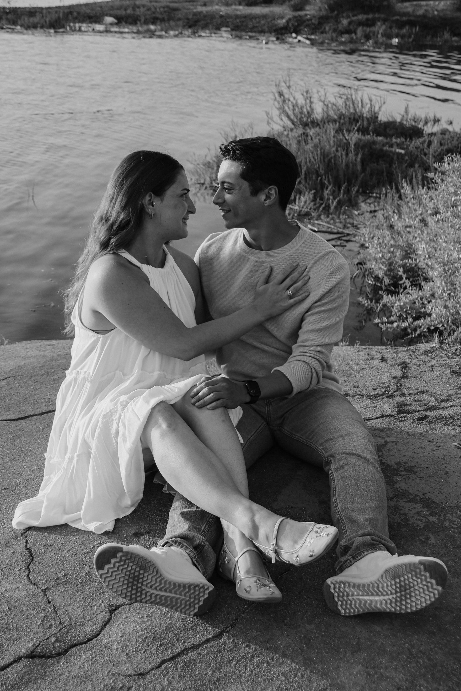 Black and white photo of couple sitting together on rocks by waterfront.