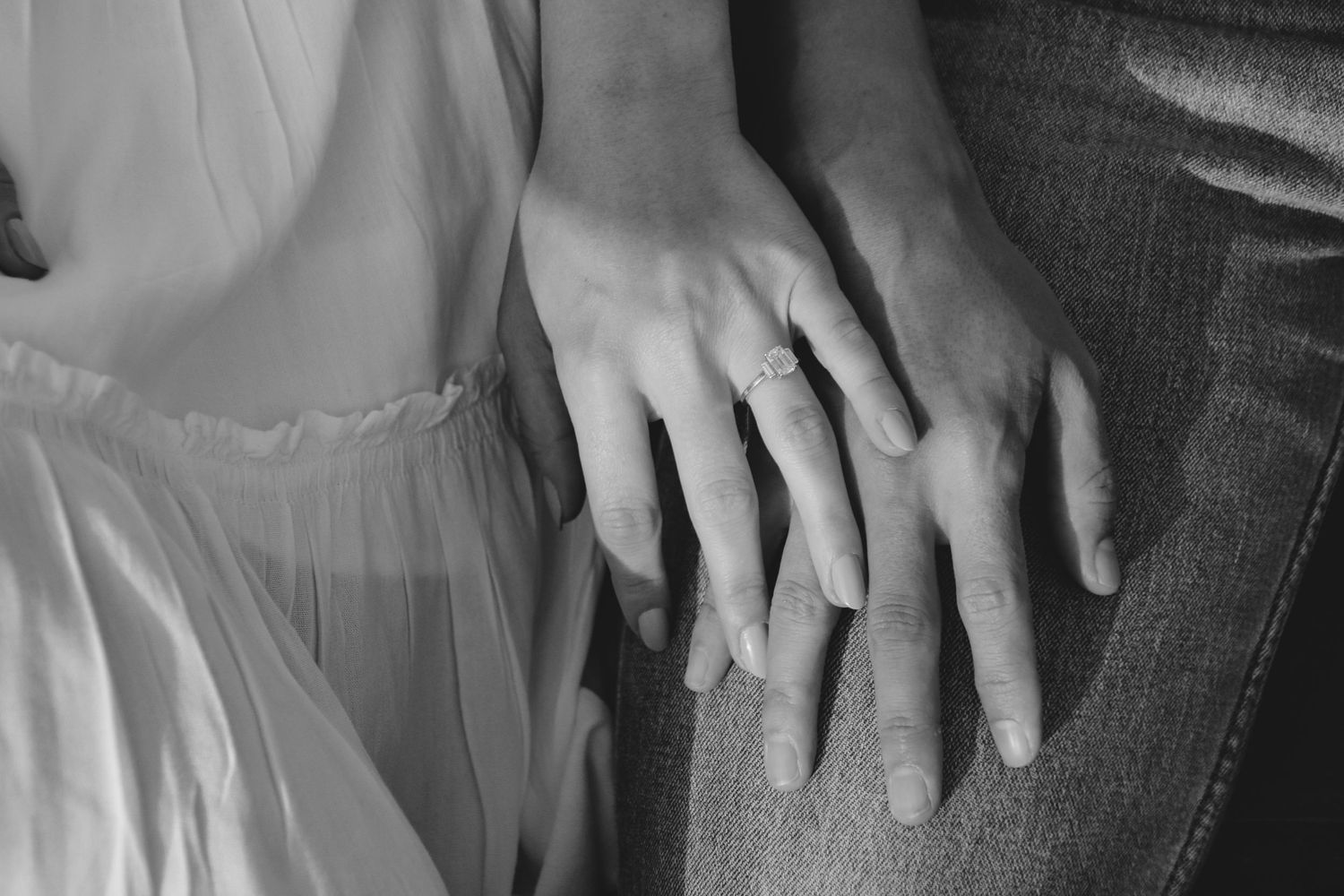 Black and white close-up of hands with engagement rings intertwined on denim and white fabric.