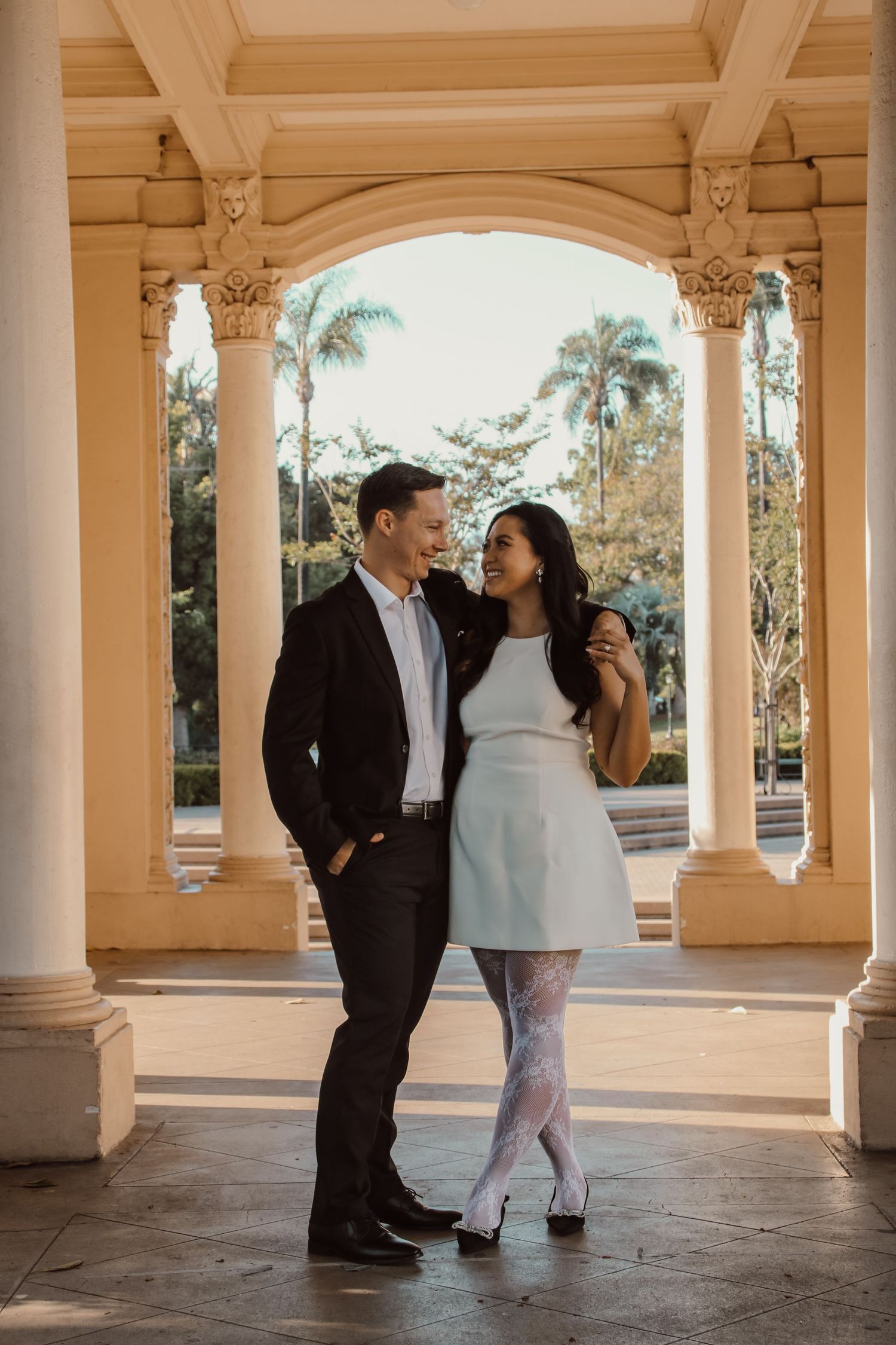 Couple sharing romantic moments under elegant columns of a beige-toned outdoor pavilion.