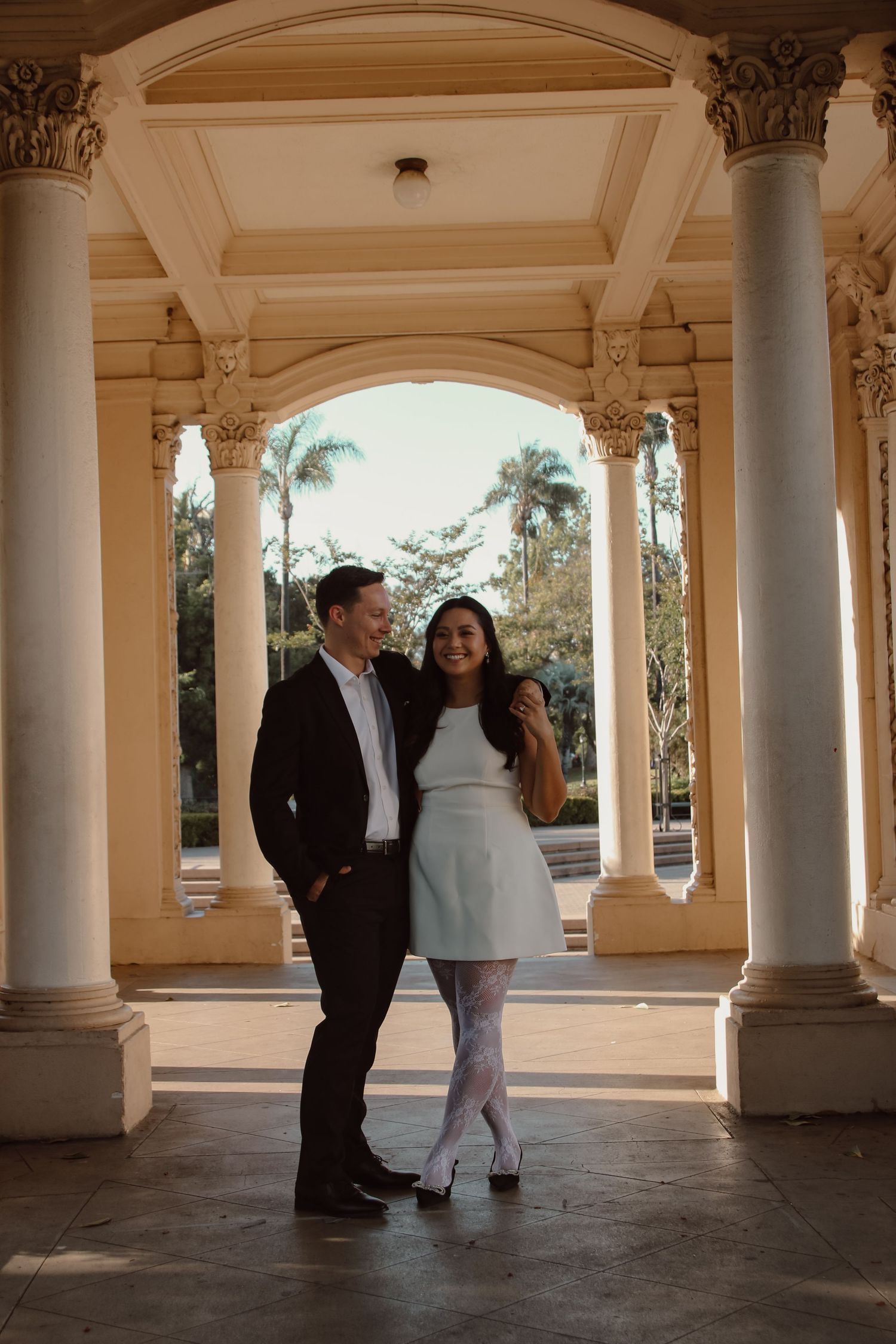 A couple laughs together in an elegant covered walkway with ornate architectural details.